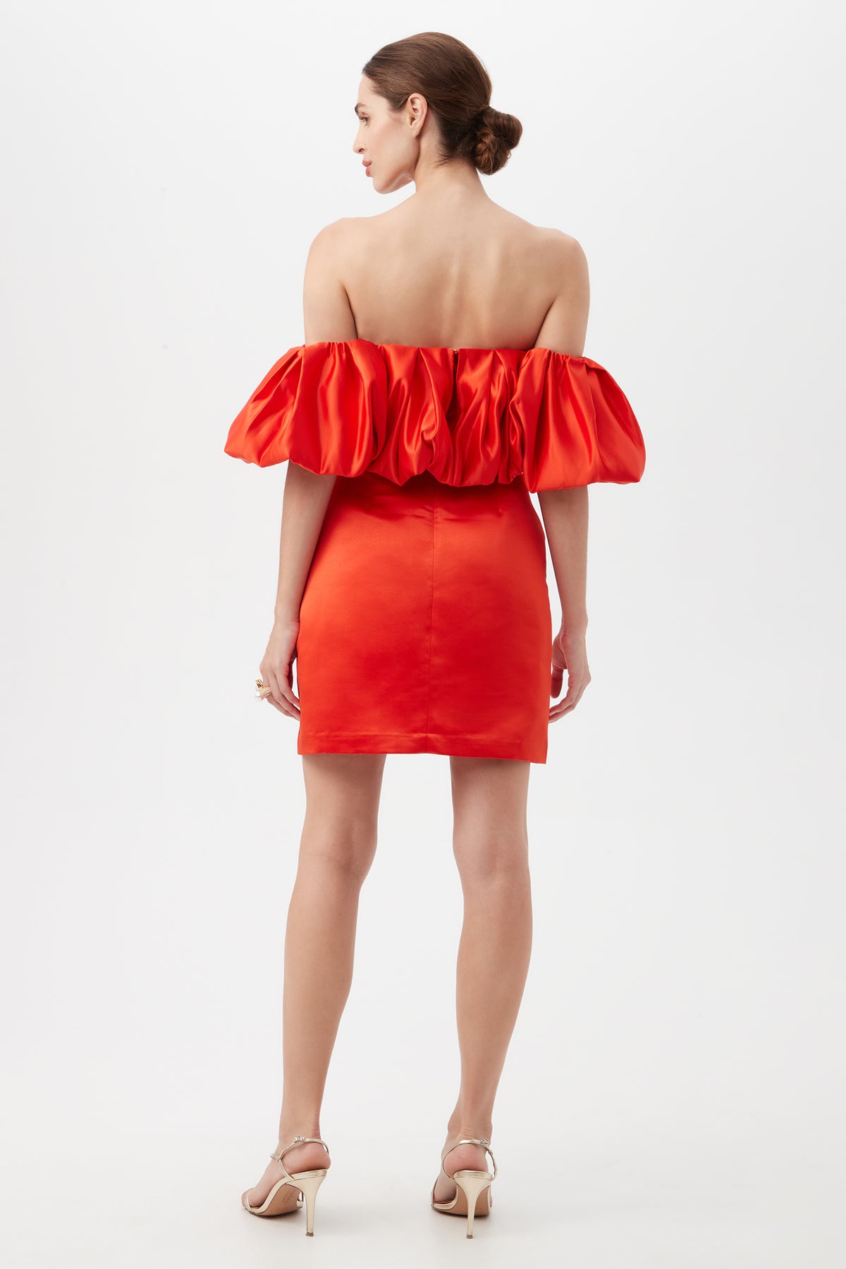 A woman faces away in the Nia Satin Off The Shoulder Short Cocktail Dress, featuring puffed sleeves and an open back. Her hair is styled in a bun, and she wears beige high-heeled sandals against a plain white background.