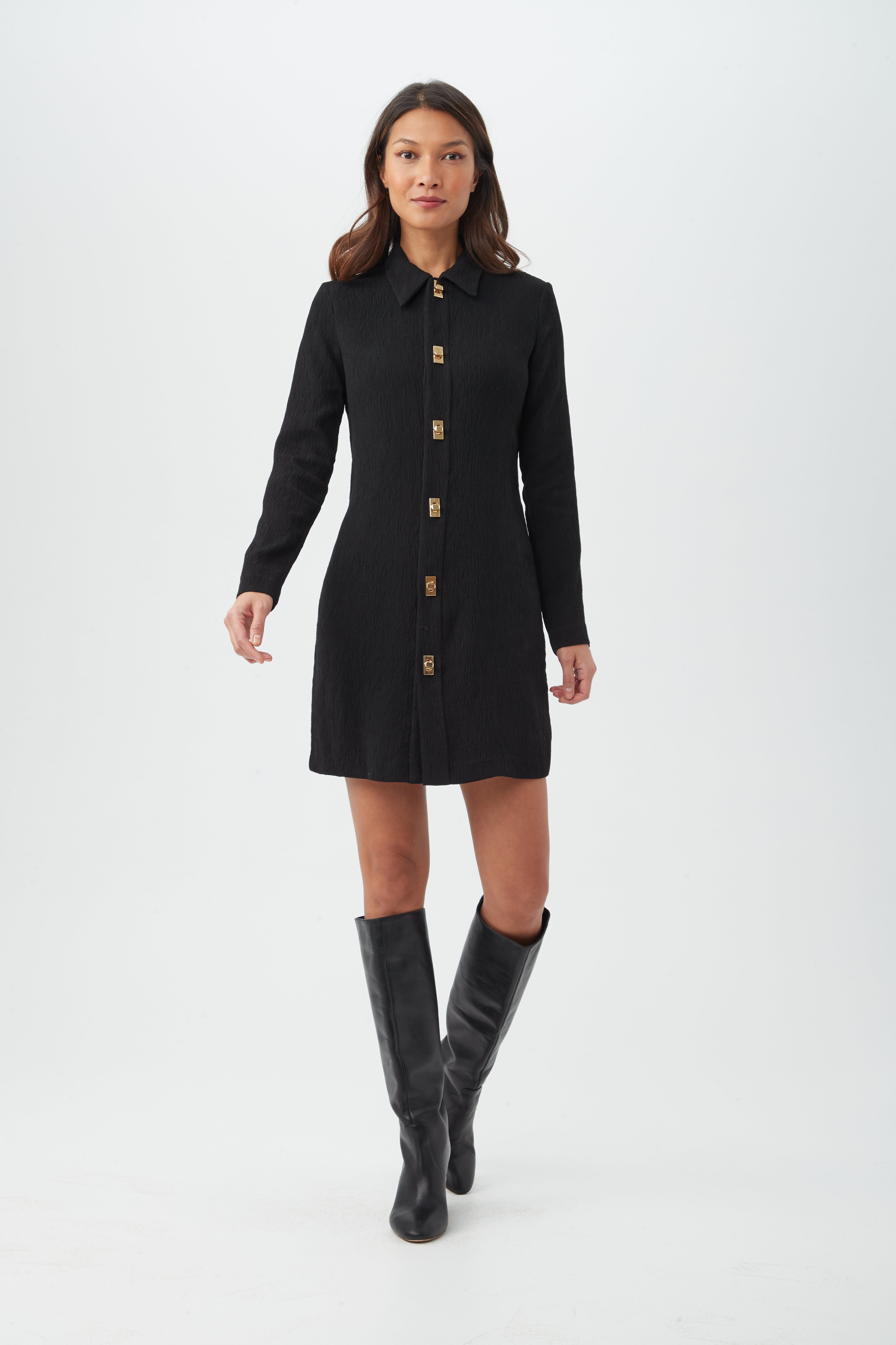 A woman wearing the Barika Dress, a long-sleeve black button-down style, and knee-high black boots walks forward against a plain white background.