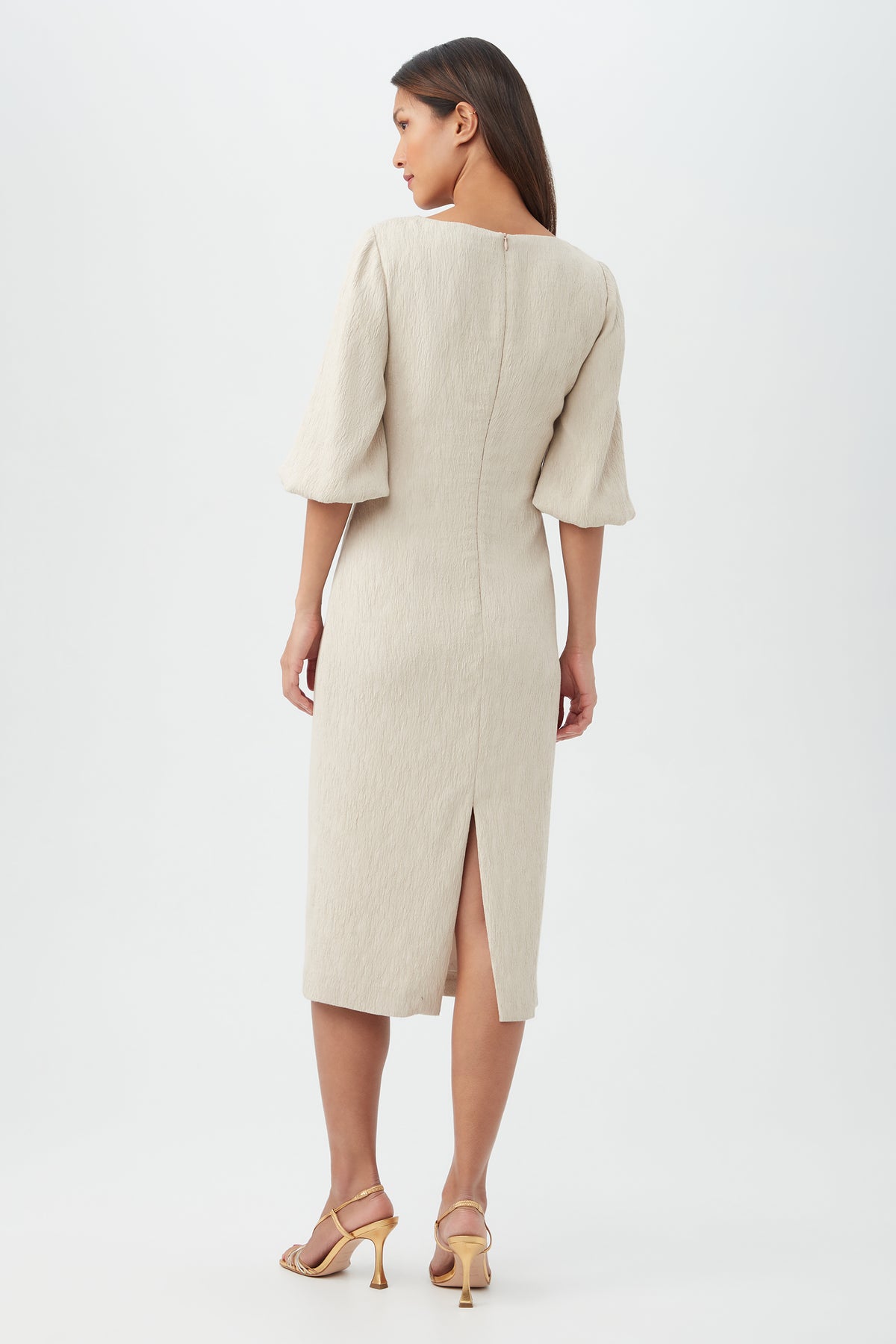 A woman stands against a plain background wearing the Madeline DressÃ¢â‚¬â€a light beige, knee-length dress with three-quarter sleeves and a back slitÃ¢â‚¬â€paired with light-colored heeled sandals. She faces away, looking to the side.