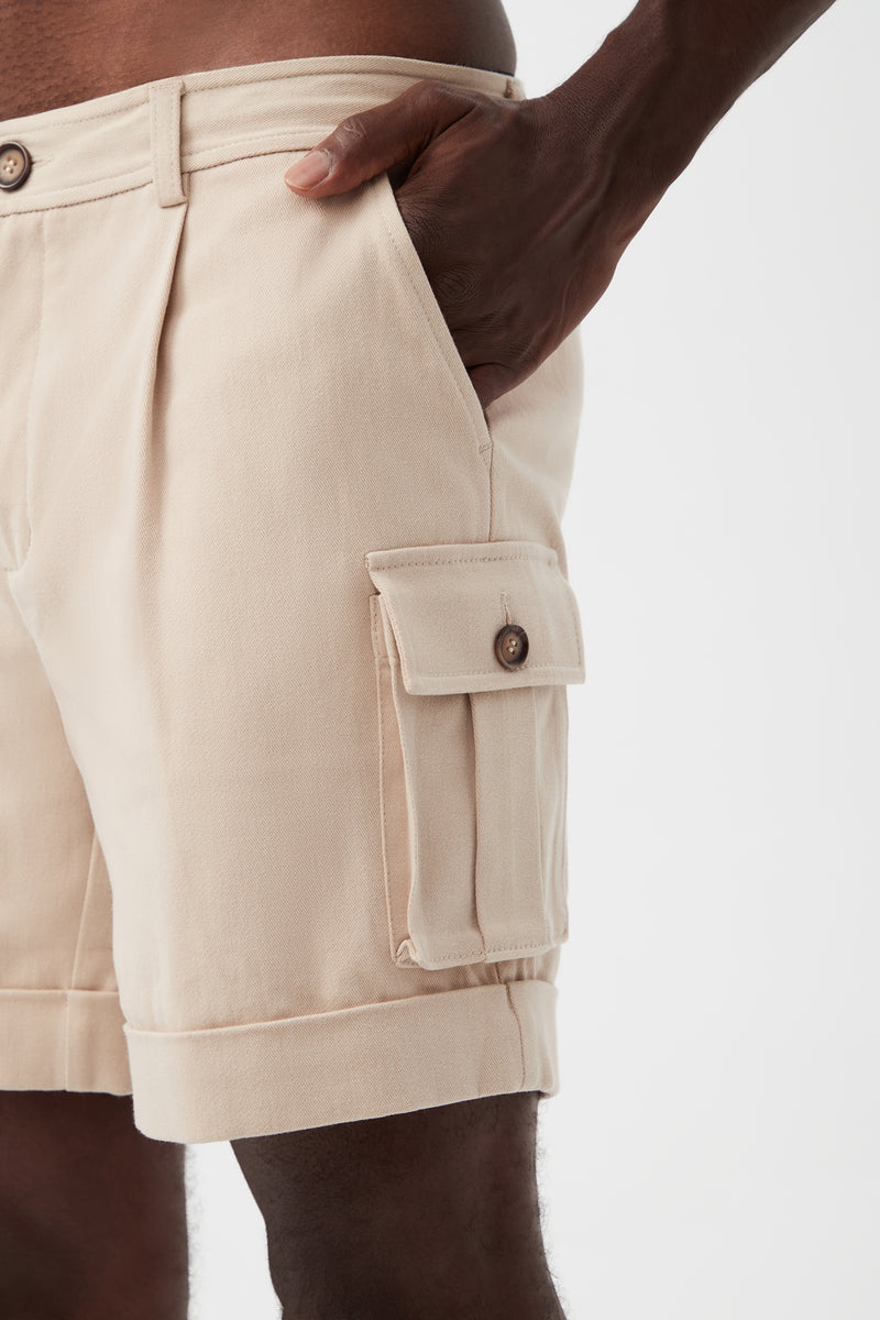 A person wears the Westlake Cargo Short in light beige brushed cotton twill, featuring a pleated front and buttoned cargo pocket, shown from waist to mid-thigh with one hand in the pocket against a plain white background.