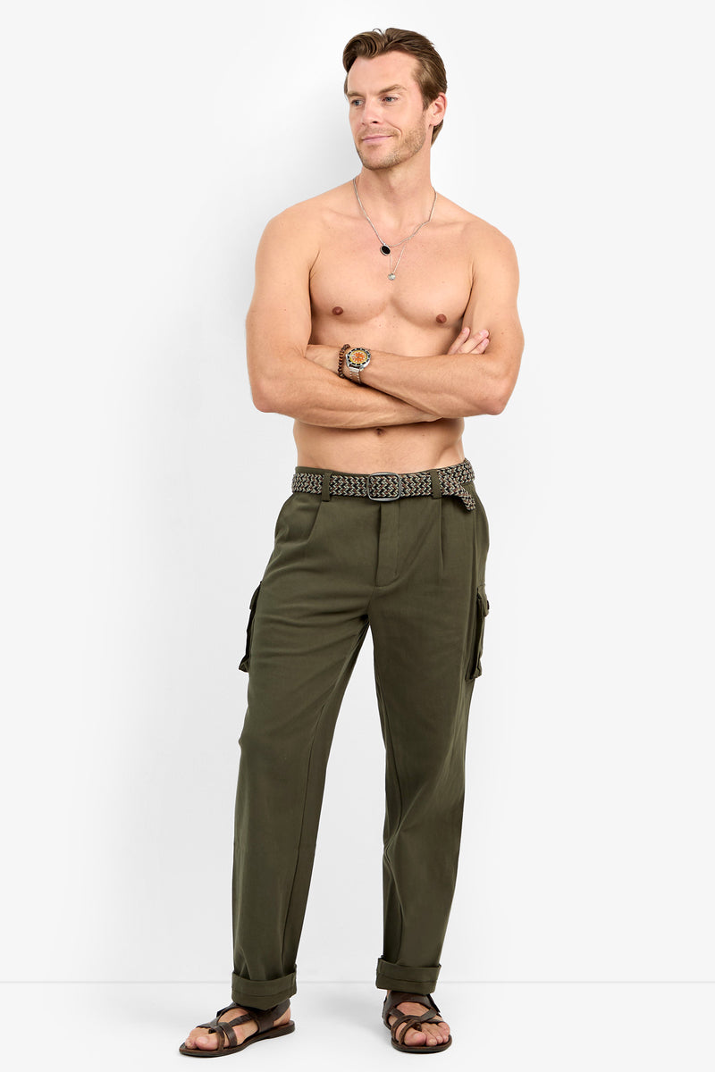A shirtless man with crossed arms wears the Lincoln Heights Cargo Trouser - Brushed Twill, a patterned belt, sandals, a watch, and a necklace, standing against a plain white background.