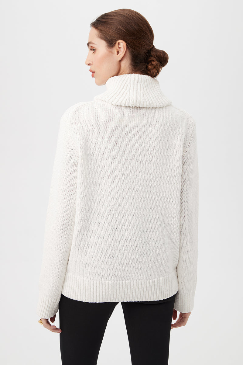 Woman with brown hair in a low bun wears the Kalahari Sweater, a white chunky knit turtleneck, paired with black pants, standing with her back to the camera against a plain light background.