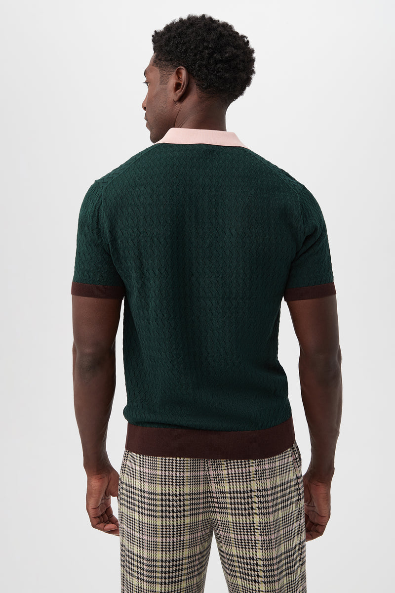 A man stands facing away, wearing the Vinny Short-Sleeve Polo in dark green with brown trim, a textured pattern, and a contrasting light collar, paired with plaid pants against a plain white background.