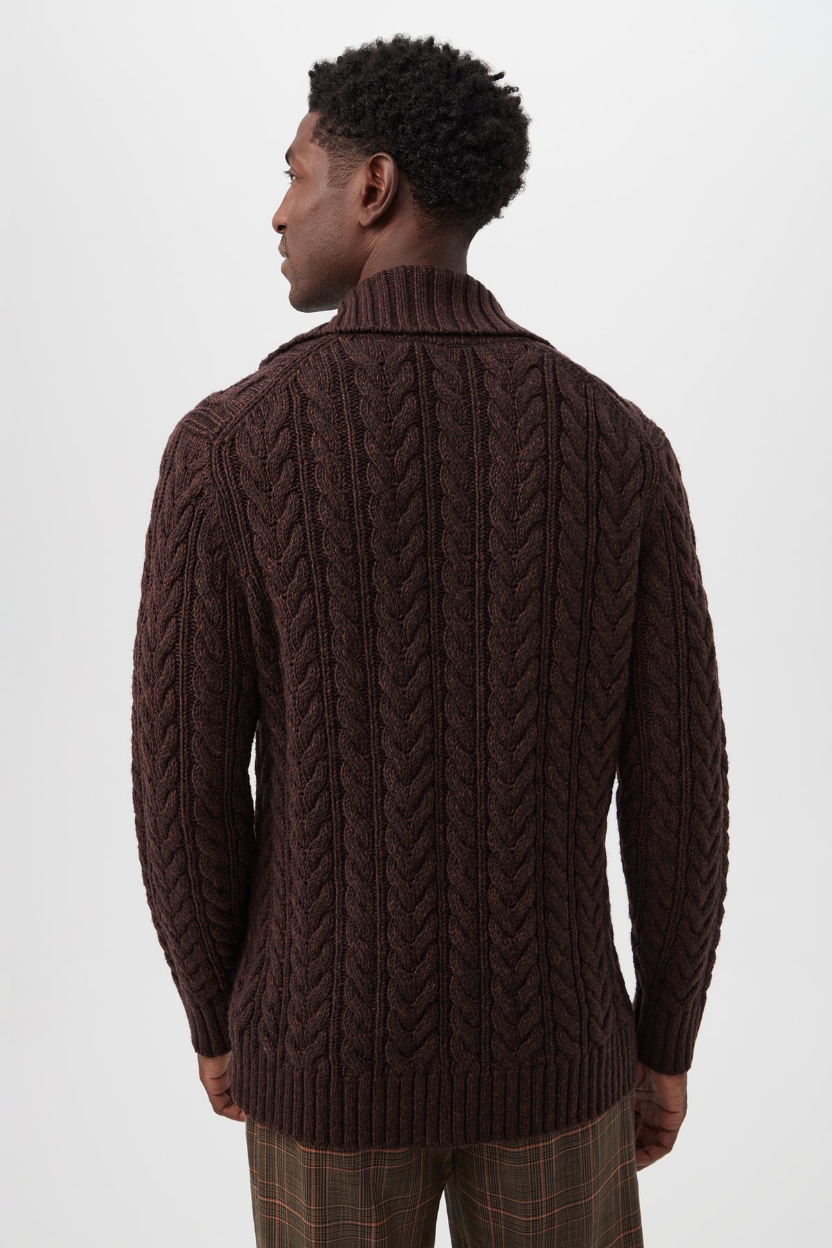 A man with short curly hair faces away, wearing the Raleigh Sweater Coat in brown merino wool featuring a chunky cable-knit and shawl collar, paired with brown plaid trousers against a plain light background.