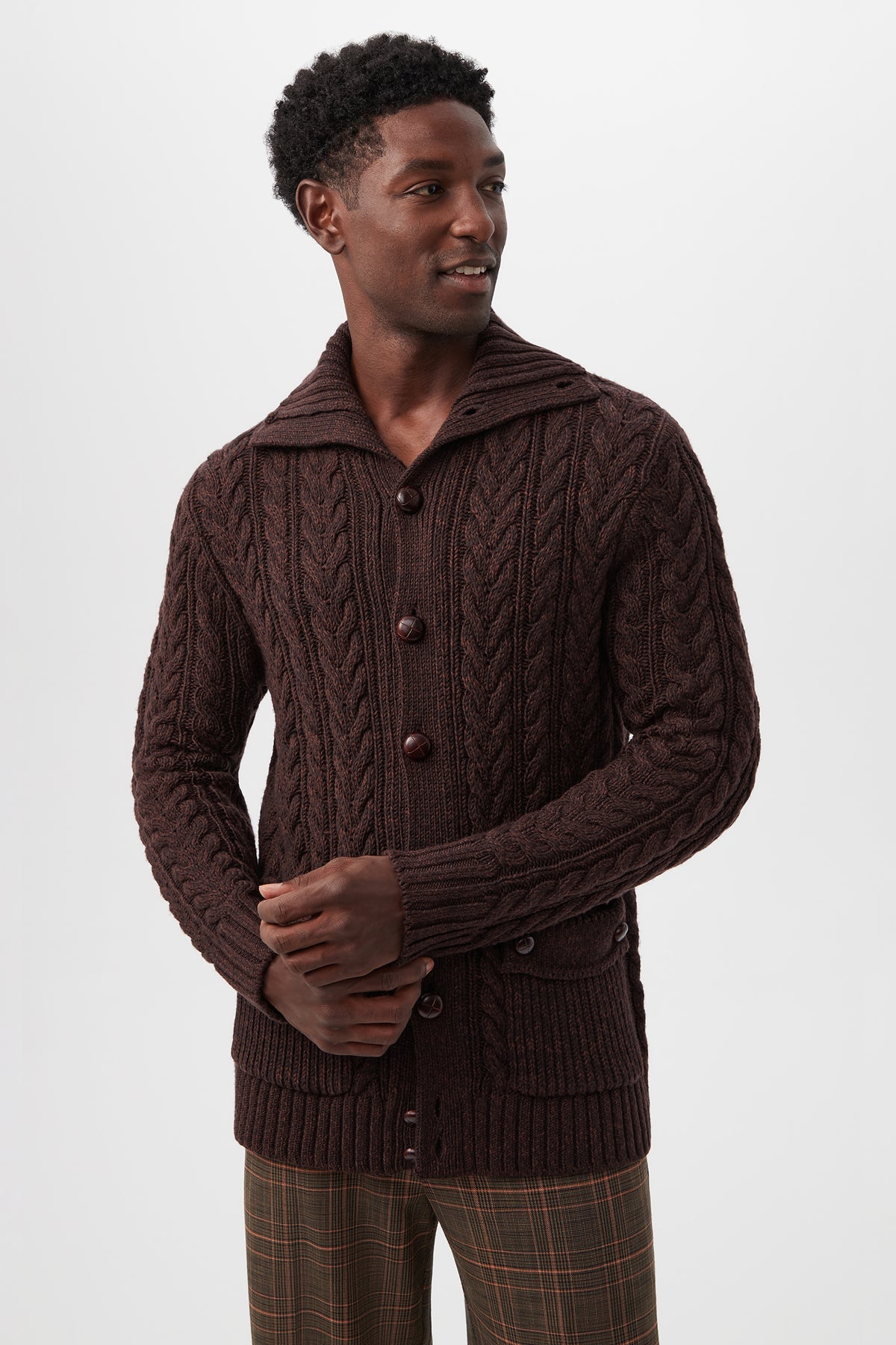 A man wearing the Raleigh Sweater Coat in Mudcloth Merino Wool, featuring a cable-knit design, large buttons, and front pockets, stands against a white background looking left with his hands loosely clasped.