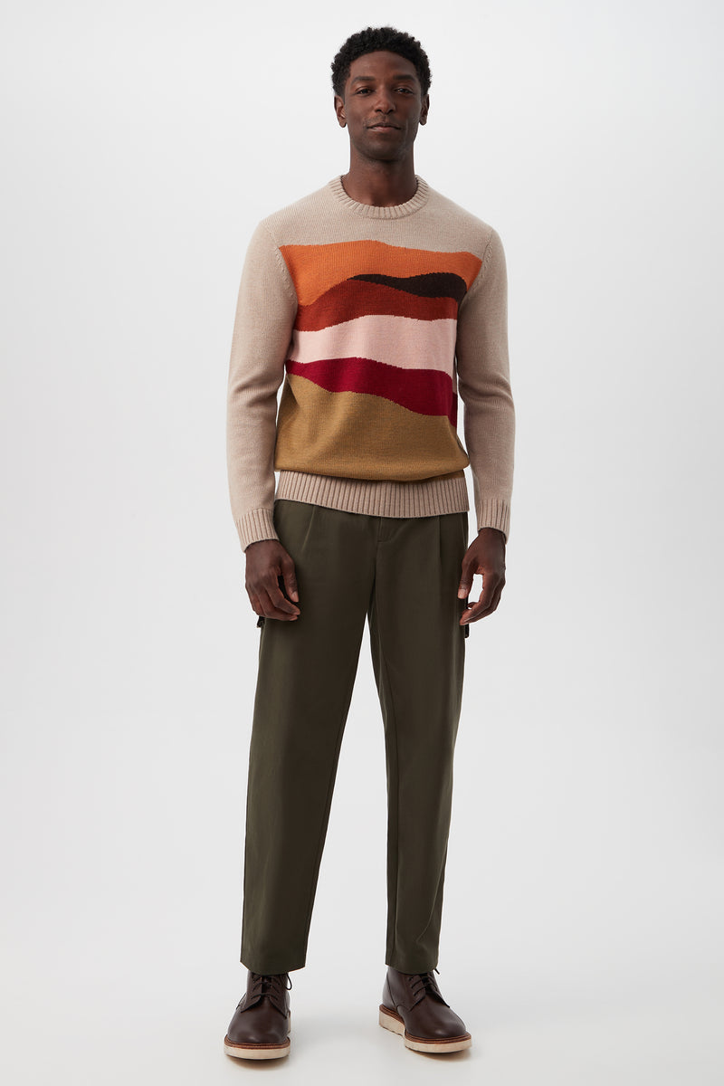 A man stands against a plain background wearing the Murphy Long-Sleeve Crewneck - Merino Wool, featuring wavy red, orange, and burgundy stripes, paired with olive green pants and brown boots. He looks relaxed with his arms at his sides.