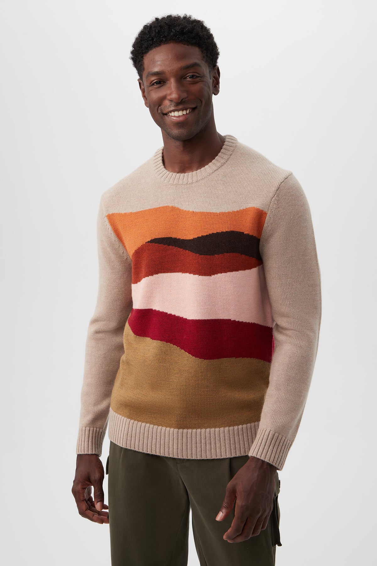 A man with short, curly hair smiles while wearing the Murphy Long-Sleeve Crewneck - Merino Wool in Camel Multi, featuring colorful wavy stripes on the chest and paired with olive green pants against a plain white background.