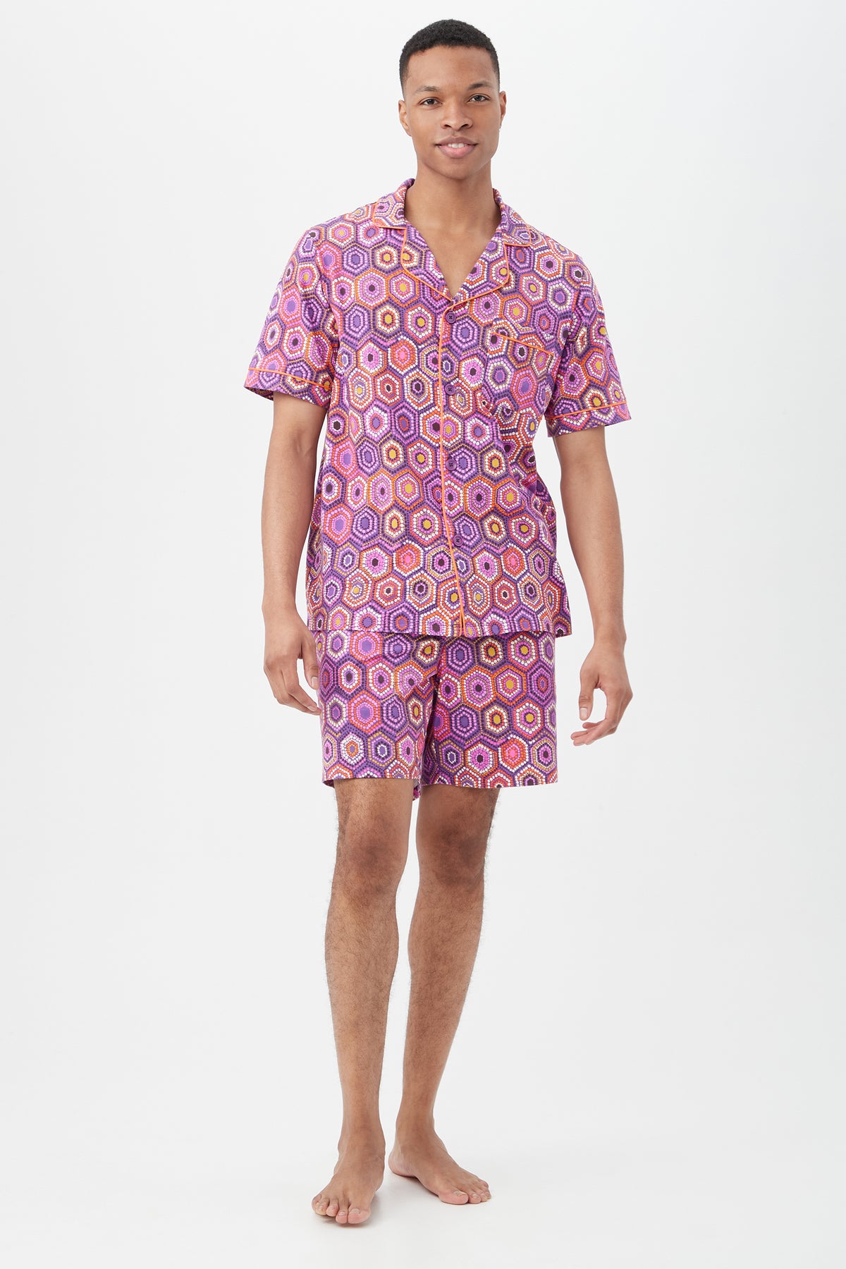 A man stands barefoot against a white background, wearing the Honeycomb Dots Mens Short Sleeve Boxer Short Jersey PJ Set featuring a colorful hexagonal pattern in purple, pink, and yellow.