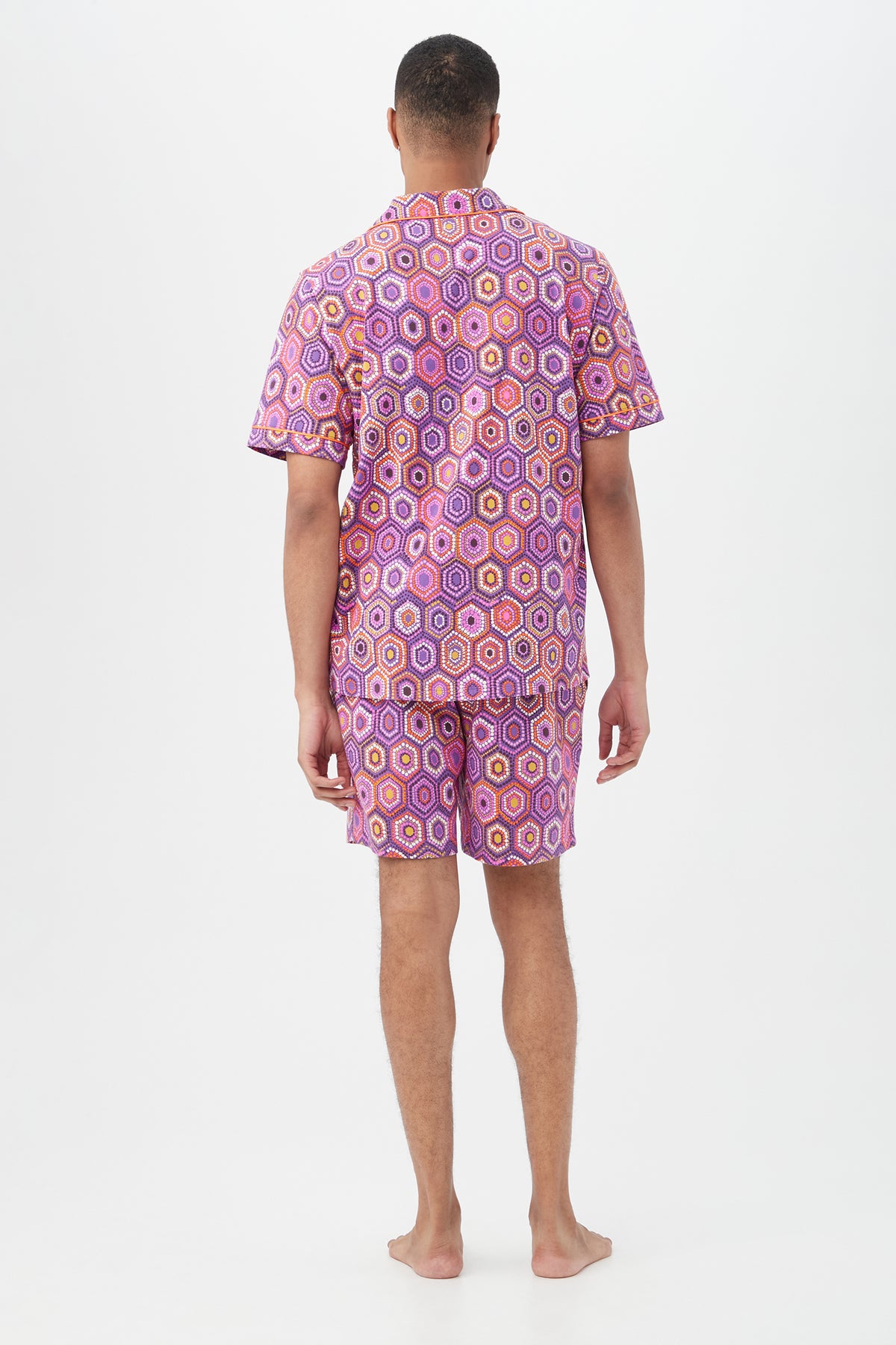 A person stands barefoot facing away, wearing the Honeycomb Dots Mens Short Sleeve Boxer Short Jersey PJ SetÃƒÂ¢Ã¢â€šÂ¬Ã¢â‚¬Âfeaturing a short sleeve button-up shirt and matching shorts in a bright pink, purple, and yellow geometric pattern on white.