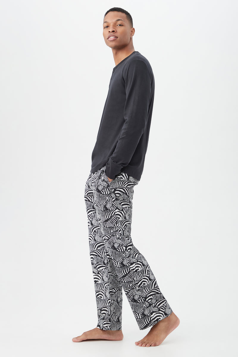 A person stands barefoot against a white background, wearing the Zebra Safari Mens Long Sleeve Henley Long Pant Poplin PJ SetÃ¢â‚¬â€featuring a black top and black-and-white abstract print pantsÃ¢â‚¬â€looking over their shoulder with hands in pockets.