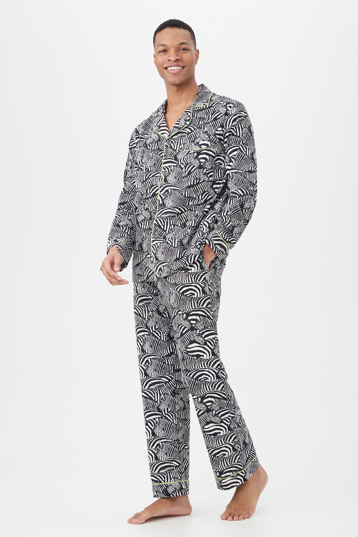 A smiling man stands barefoot against a plain white background, wearing the Zebra Safari Mens Long Sleeve Long Pant Poplin PJ Set, featuring a black-and-white zebra print with buttons and pockets in lightweight sleepwear fabric.