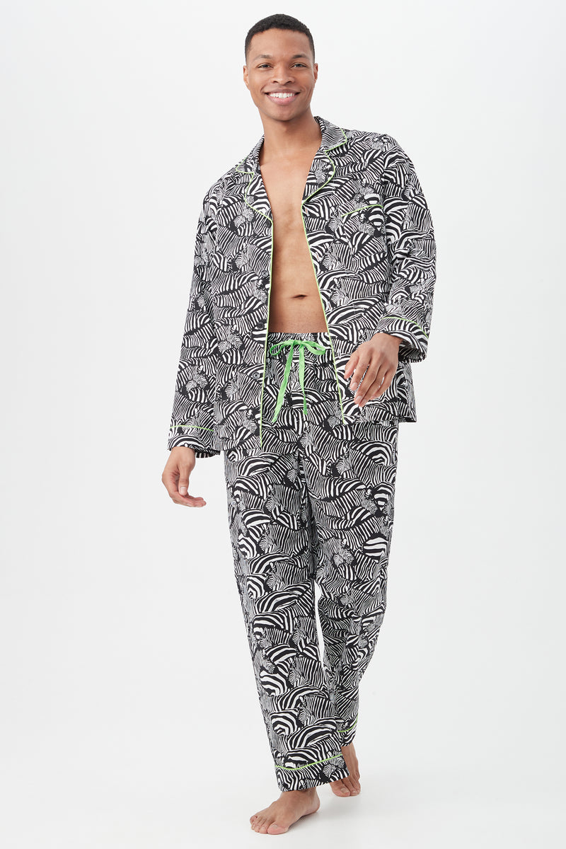 A smiling person walks barefoot in the Zebra Safari Mens Long Sleeve Long Pant Poplin PJ Setâ€”featuring a black-and-white pattern with green trim and drawstring. The lightweight sleepwear top is open, revealing their chest, against a plain white background.