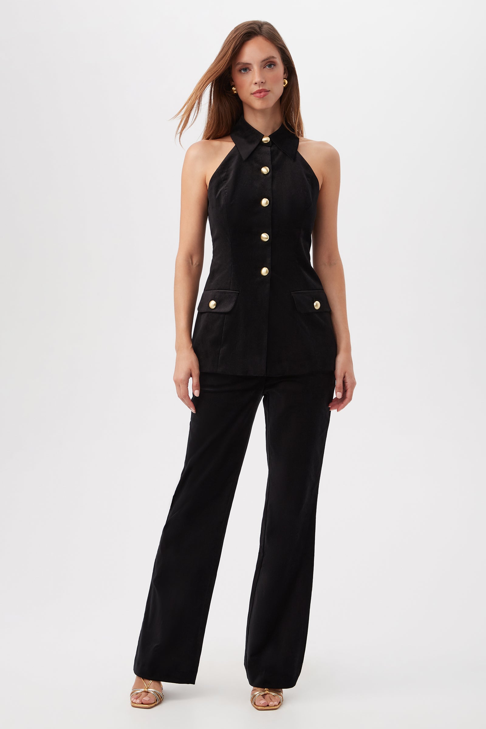 A woman with long brown hair poses against a white background in the Elainia Velveteen Sleeveless Tunic Top, a black, collared button-up featuring gold buttons. She pairs it with matching black pants and gold sandals.