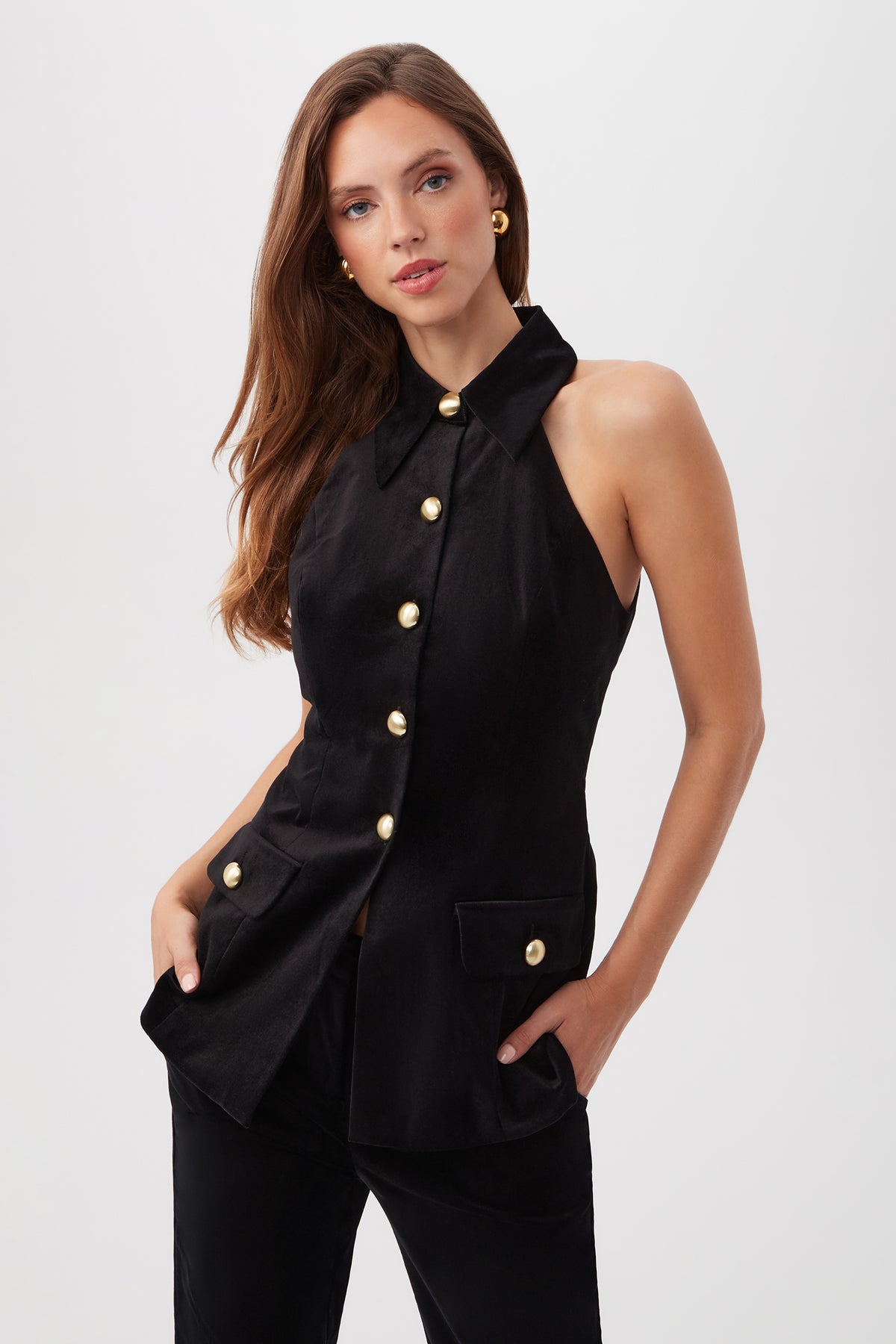 A woman in the Elainia Velveteen Sleeveless Tunic Top in black, paired with gold hoop earrings and black pants, poses against a plain white background with one hand in her pocket.