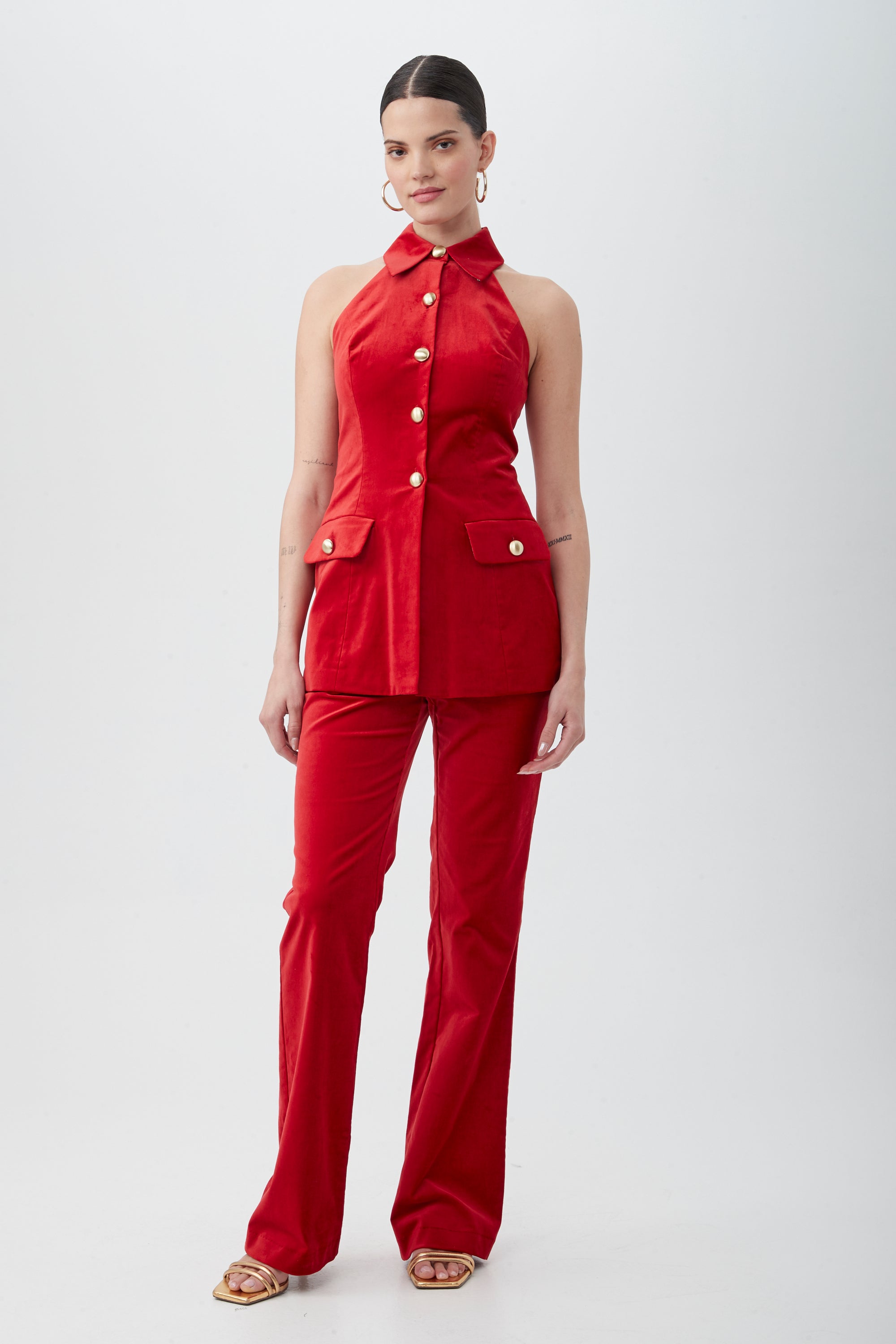 A woman stands against a white background wearing the Elainia Velveteen Sleeveless Tunic Top in red with a collar and matching pants. Her hair is tied back, and she accessorizes with hoop earrings and open-toe sandals.