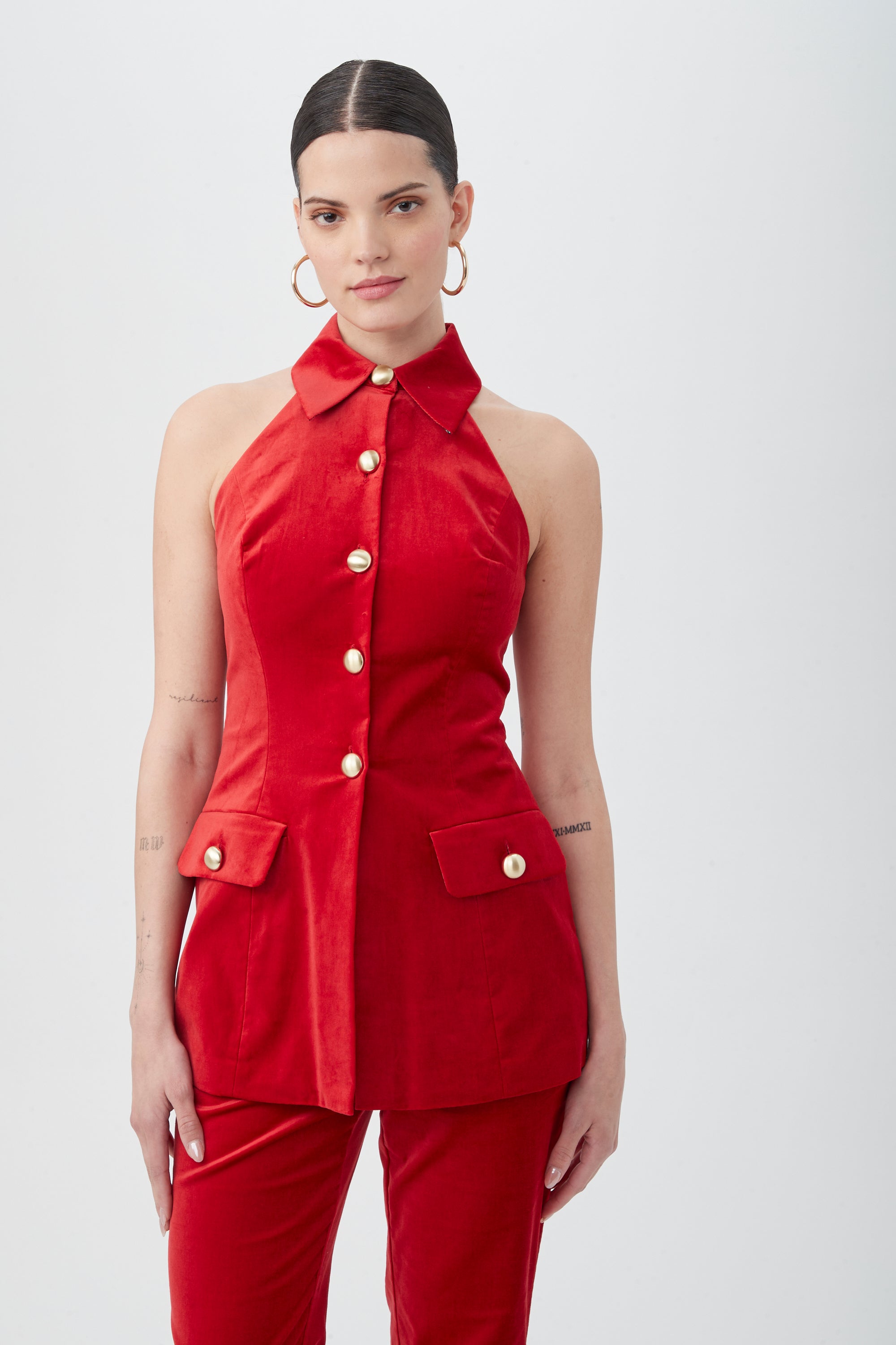 A woman wears the Elainia Velveteen Sleeveless Tunic Top in red, featuring gold buttons and flap pockets, with matching red pants. She has slicked-back hair, gold hoop earrings, and neutral makeup against a plain white background.