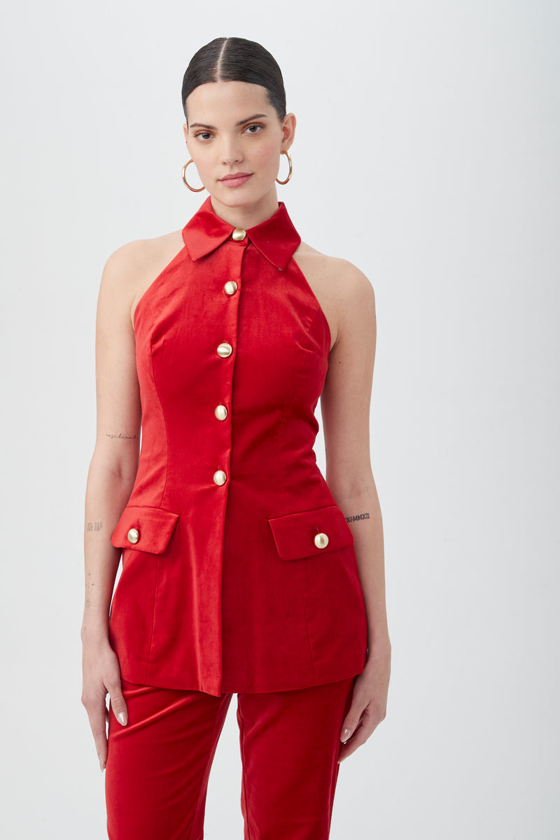 A woman wears the Elainia Velveteen Sleeveless Tunic Top in red, featuring gold buttons and flap pockets, with matching red pants. She has slicked-back hair, gold hoop earrings, and neutral makeup against a plain white background.