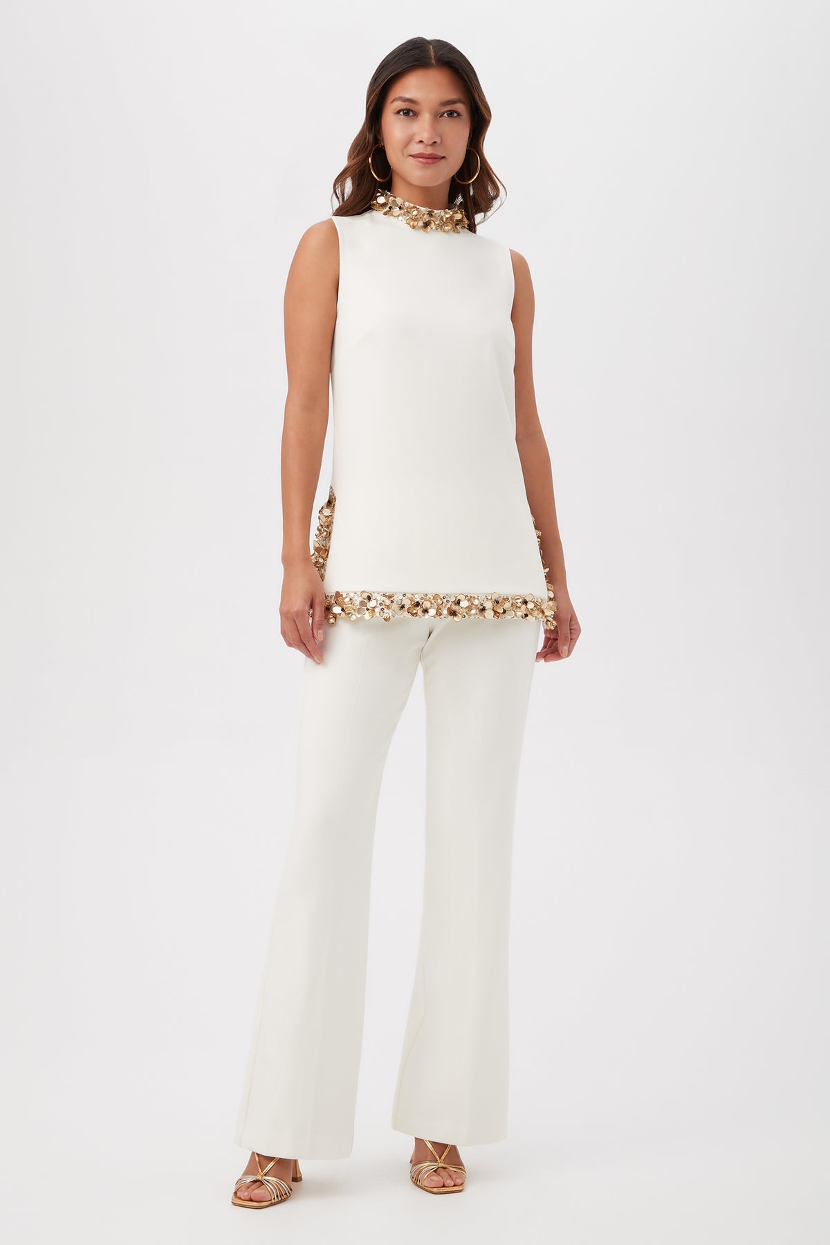 A woman wears the Wonderous 2 Sleeveless Mockneck Tunic Top With Gold Beading, styled with matching white flared pants and gold strappy heels, standing against a plain white background.