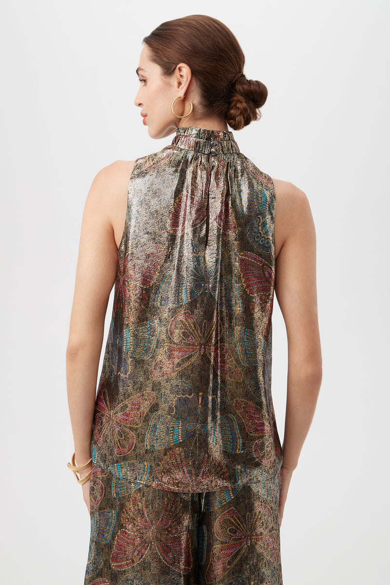 A woman with brown hair in a low bun is seen from the back, wearing the Rissa Butterfly Print Silk Sleeveless Mockneck Top featuring a metallic, multicolored butterfly design. She accessorizes with gold hoop earrings and bracelets.