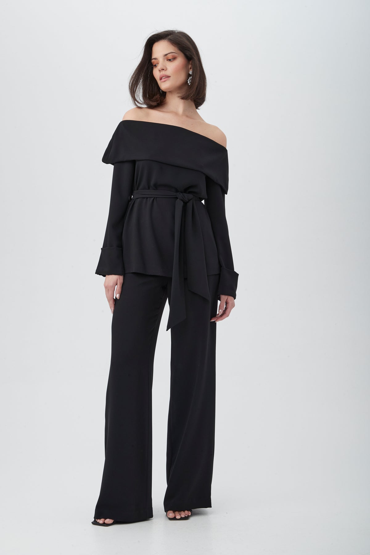 A woman wears the Lyndi Off-The-Shoulder Belted Long Sleeve Top in black with matching wide-leg pants and open-toe shoes, standing against a plain white background.