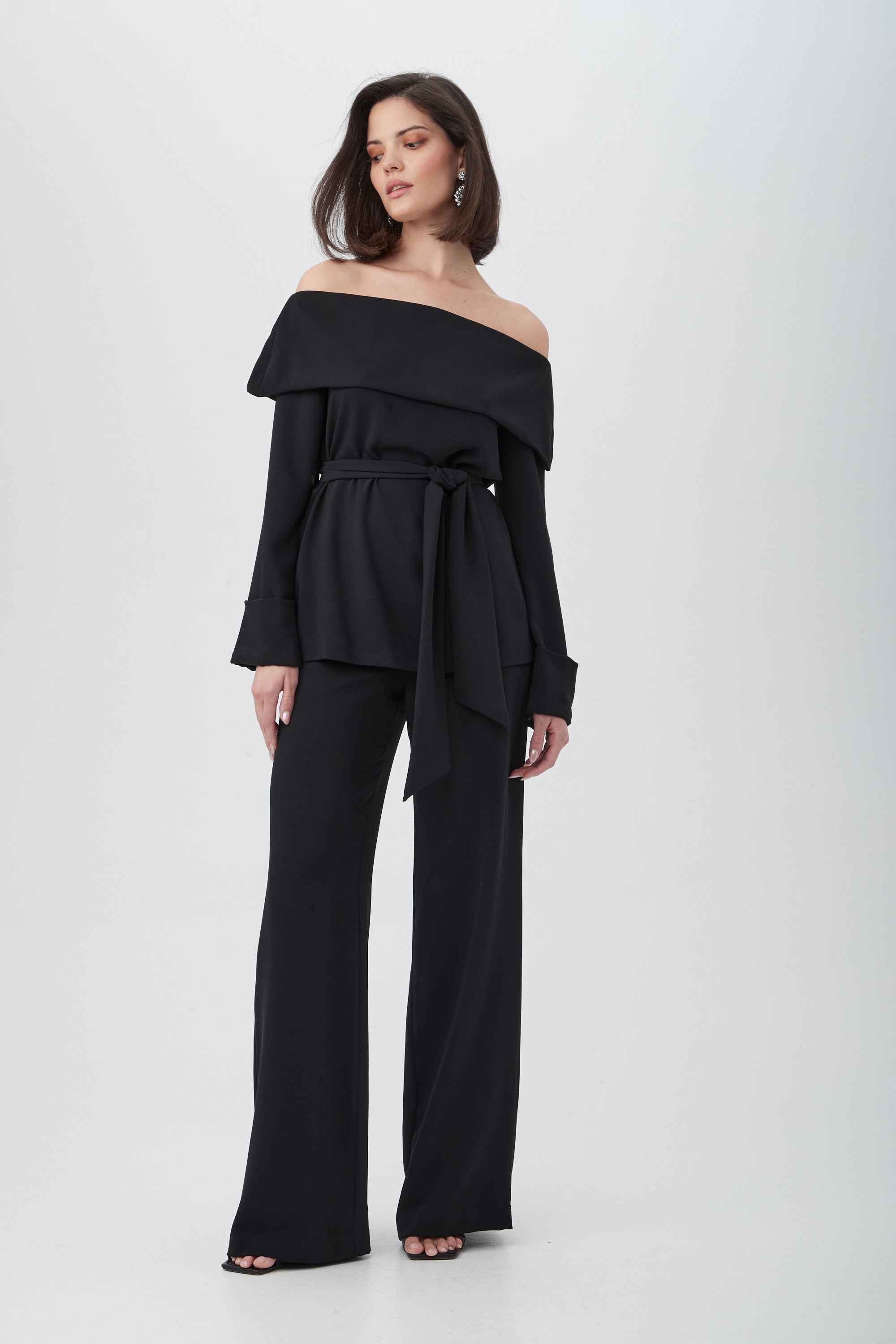A woman wears the Lyndi Off-The-Shoulder Belted Long Sleeve Top in black with matching wide-leg pants and open-toe shoes, standing against a plain white background.