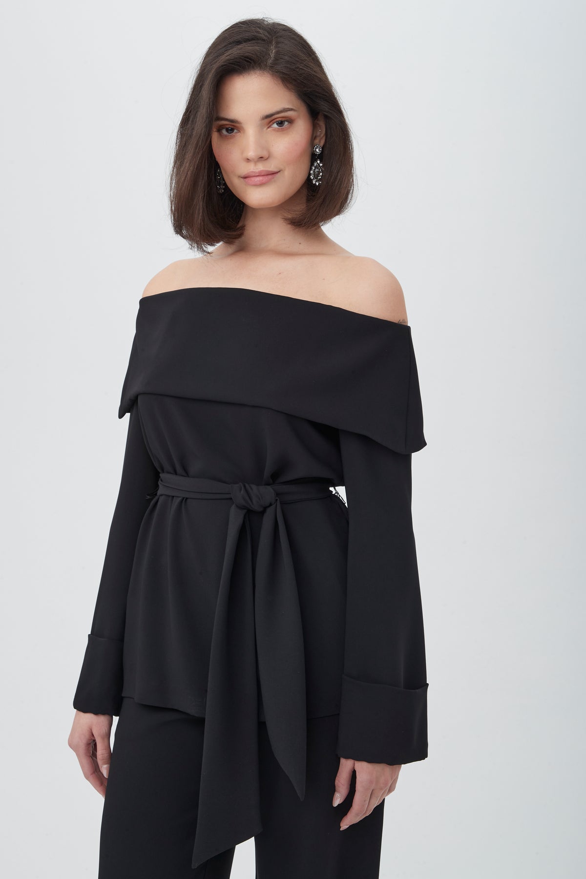 A woman with shoulder-length dark hair smiles softly as she wears the Lyndi Off-The-Shoulder Belted Long Sleeve Top in black, paired with black pants and statement earrings. The background is plain white.