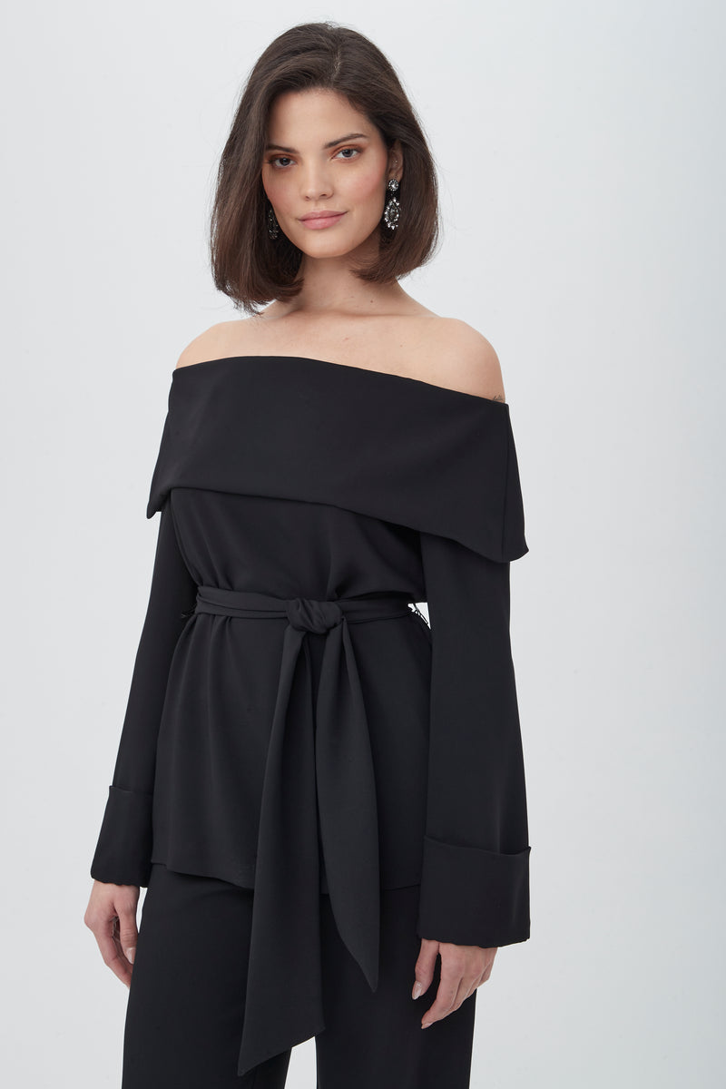 A woman with shoulder-length dark hair smiles softly as she wears the Lyndi Off-The-Shoulder Belted Long Sleeve Top in black, paired with black pants and statement earrings. The background is plain white.