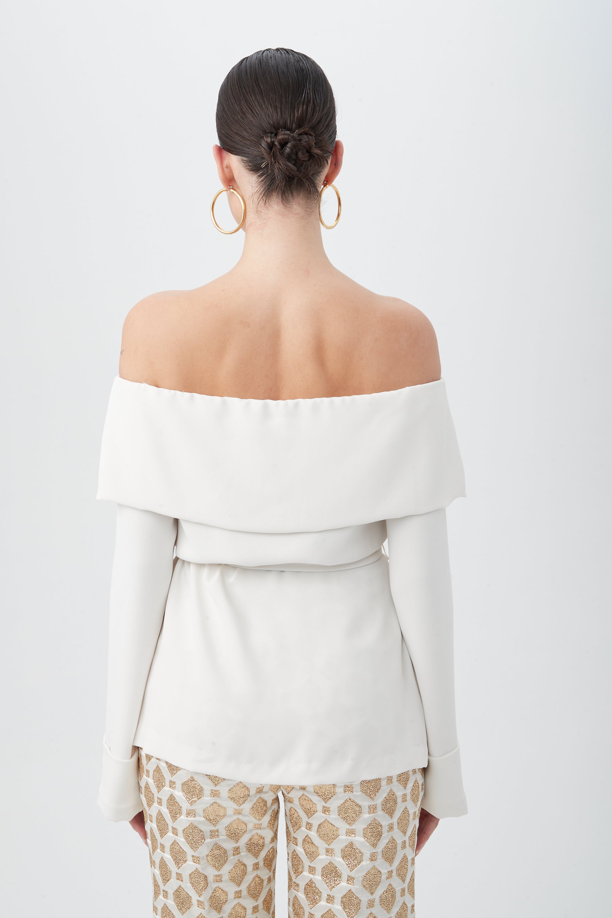 A woman with dark hair in a low bun wears the Lyndi Off-The-Shoulder Belted Long Sleeve Top, paired with patterned beige pants and gold hoop earrings, standing against a plain white background.