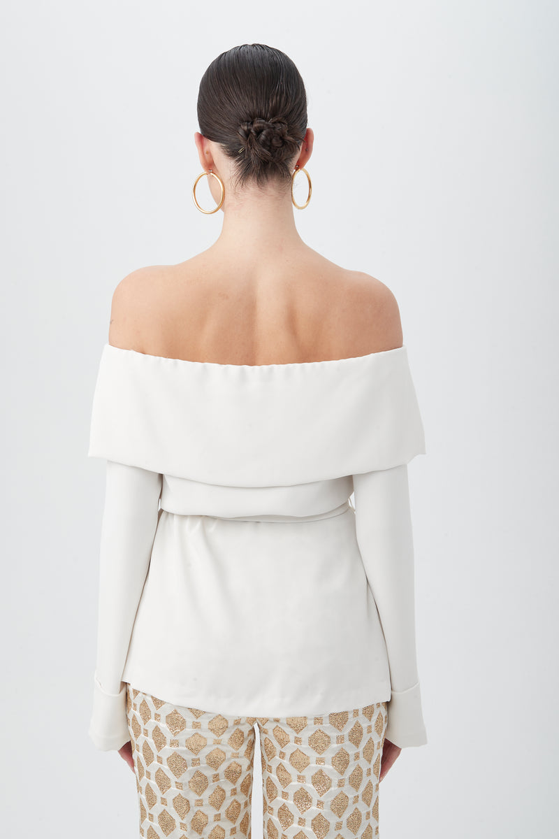A woman with dark hair in a low bun wears the Lyndi Off-The-Shoulder Belted Long Sleeve Top, paired with patterned beige pants and gold hoop earrings, standing against a plain white background.