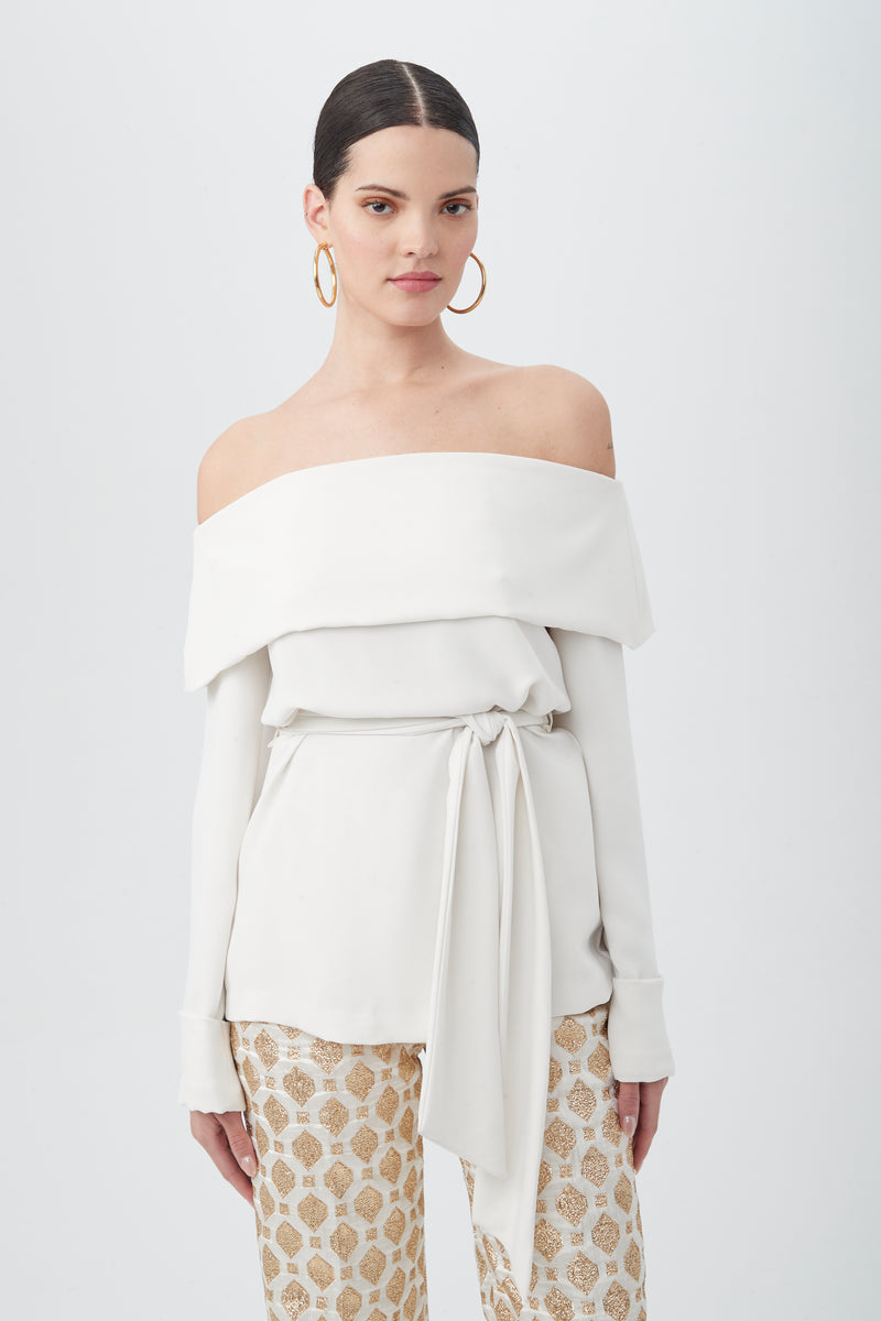 A woman wears the Lyndi Off-The-Shoulder Belted Long Sleeve Top in white with patterned beige-and-white pants, standing against a plain white background.