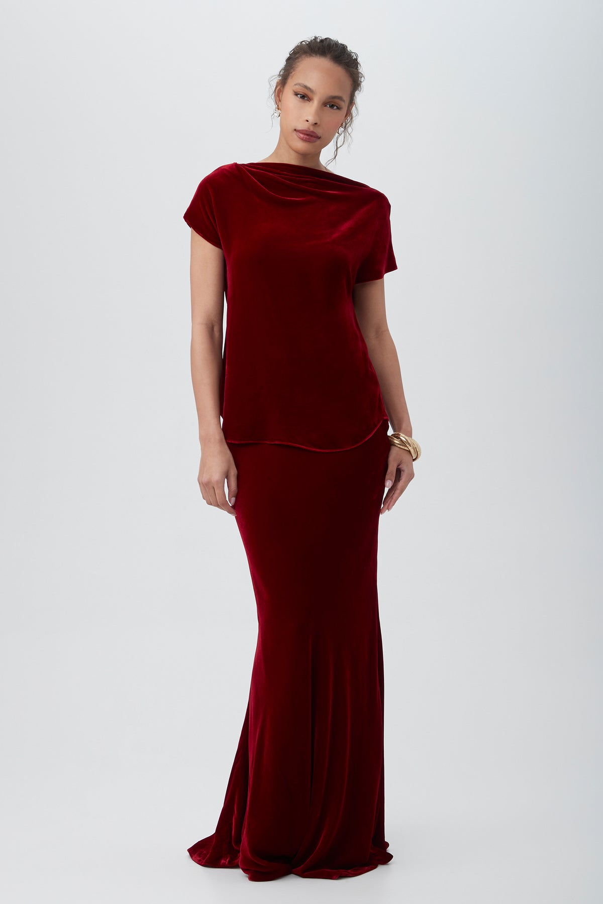 A woman wears the Sintha Velvet Boatneck Short Sleeve Top in deep red, styled with her hair pulled back and a gold bracelet, standing against a plain light background.