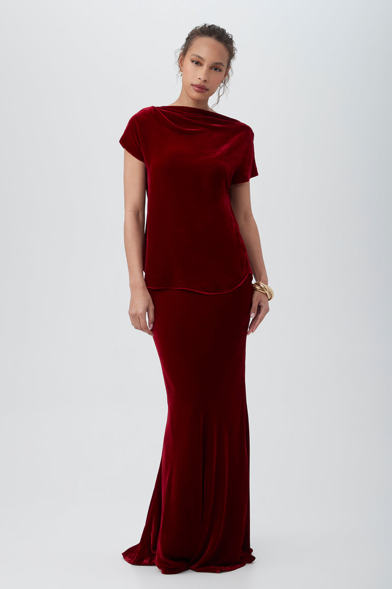 A woman wears the Sintha Velvet Boatneck Short Sleeve Top in deep red, styled with her hair pulled back and a gold bracelet, standing against a plain light background.