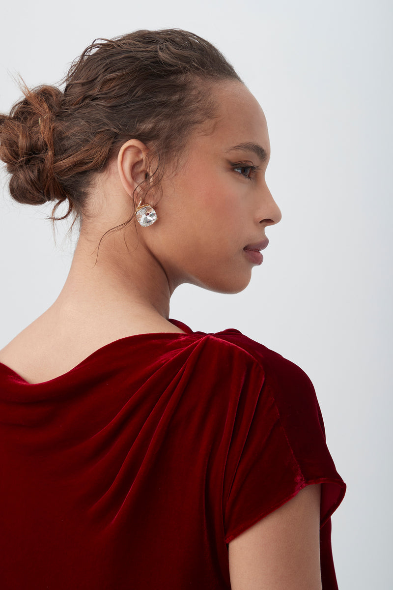 A woman with brown hair in a low bun wears the Sintha Velvet Boatneck Short Sleeve Top in deep red and large, round, sparkly earrings. She is turned slightly to the side, looking right against a plain light background.
