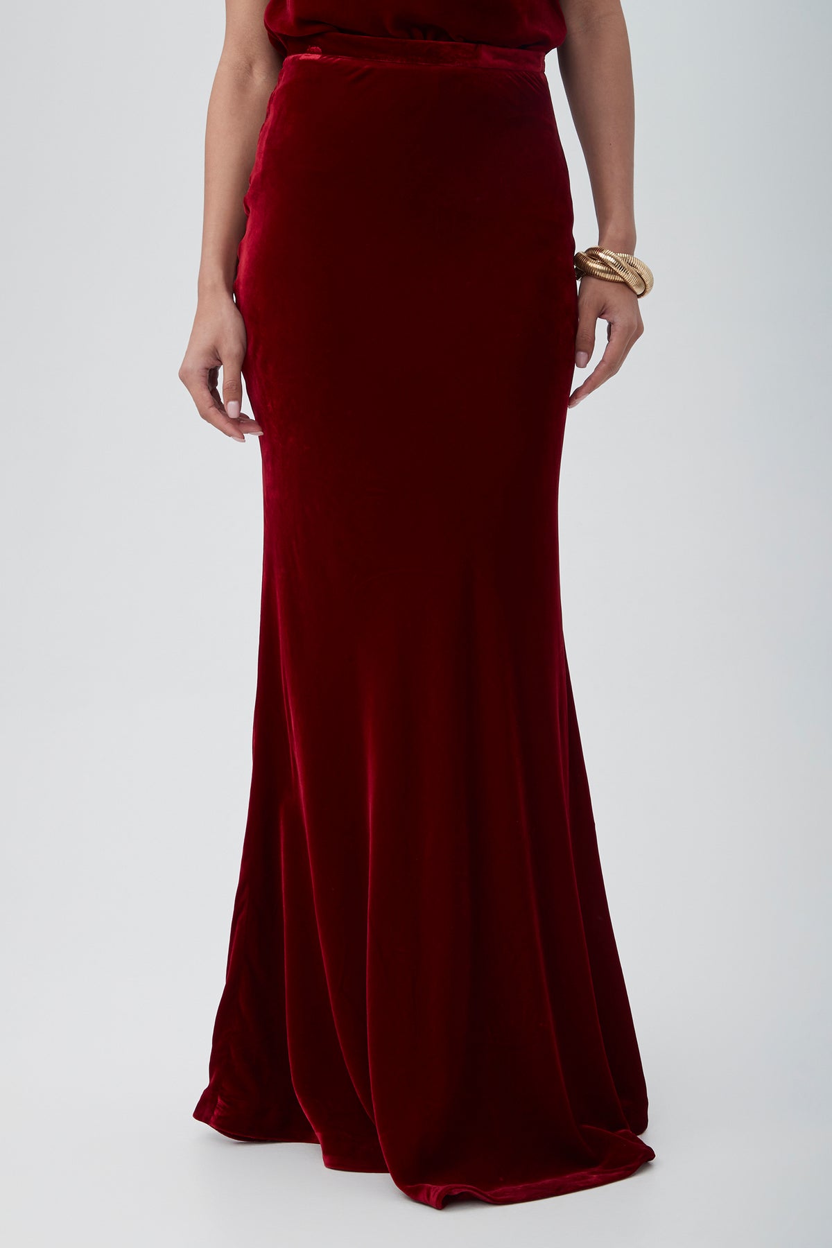 Wearing the Rabina Velvet Mermaid Maxi Skirt in deep red and a gold bracelet on their right wrist, a person stands against a plain light background.