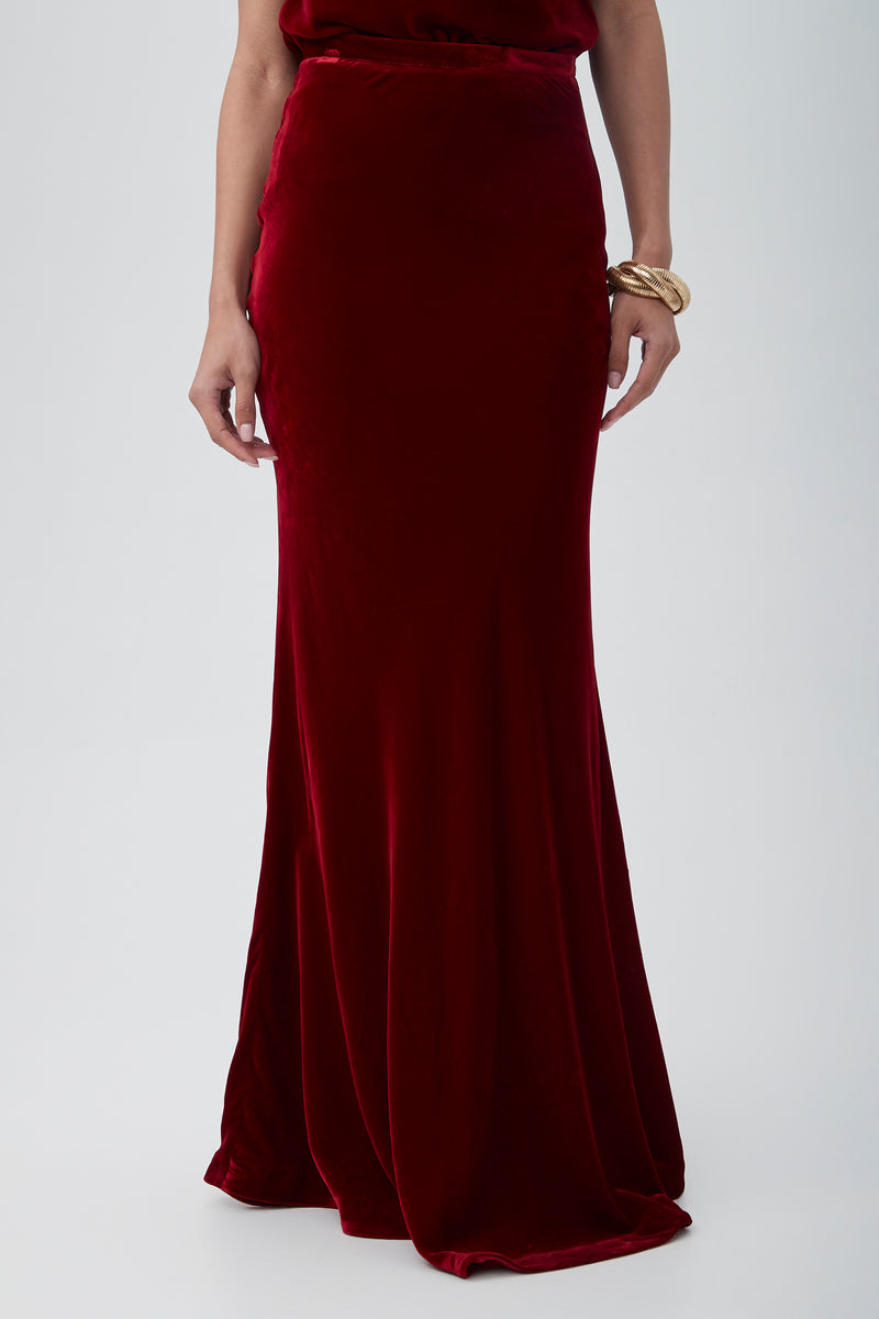 Wearing the Rabina Velvet Mermaid Maxi Skirt in deep red and a gold bracelet on their right wrist, a person stands against a plain light background.