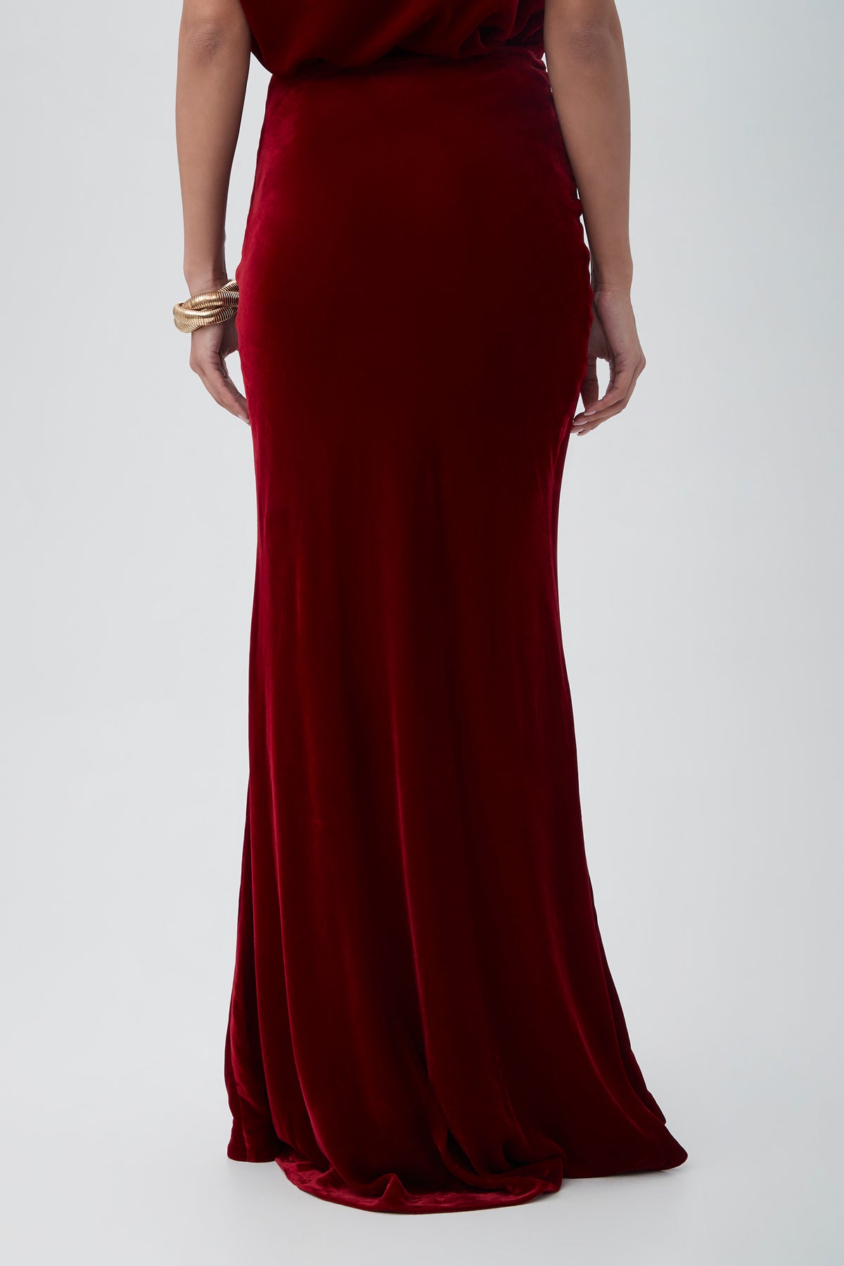 A person wearing the Rabina Velvet Mermaid Maxi Skirt in Vixen Red, shown from the back with bare arms and several gold bangles on one wrist, stands against a plain light background.