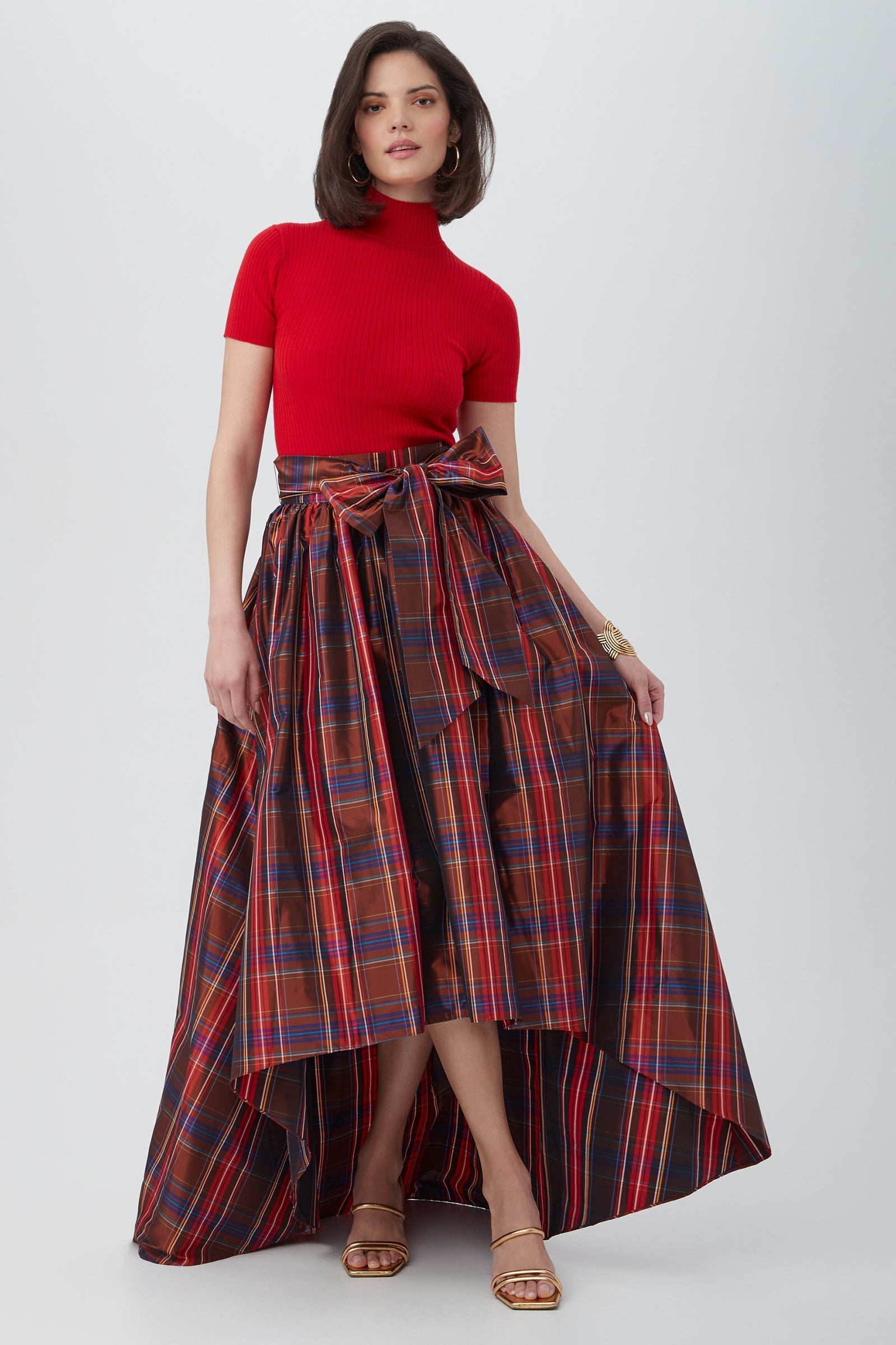 A woman poses against a plain light background wearing the Armeni Merino Wool Short Sleeve Mockneck Top in red, paired with a high-low red plaid skirt with a large waist bow and gold sandals.