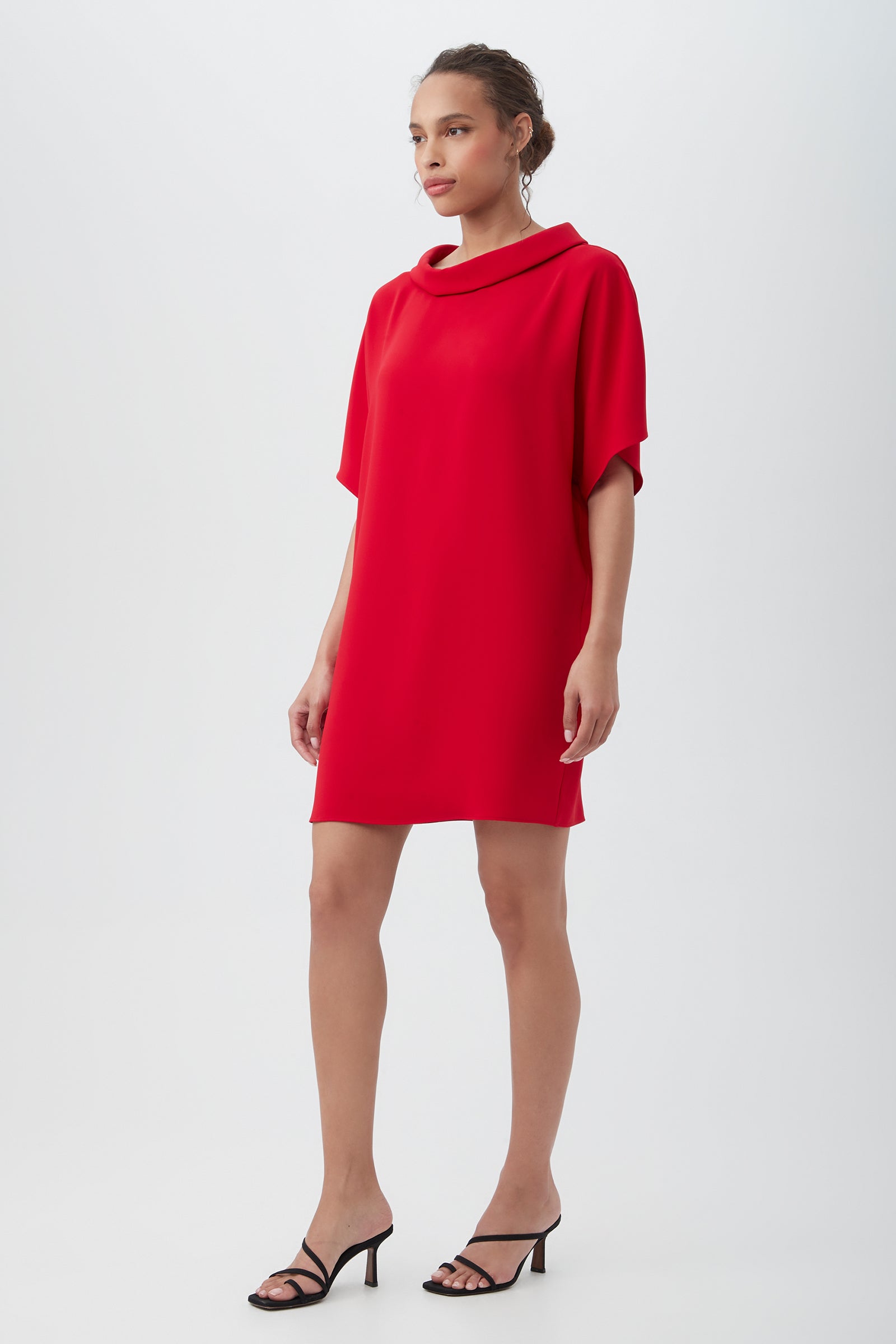 A woman stands in a simple studio wearing the Honeysuckle Short Dolman Sleeve Shift Dress—a loose, knee-length red dress with short sleeves and a draped neckline—paired with black strappy high-heeled sandals.