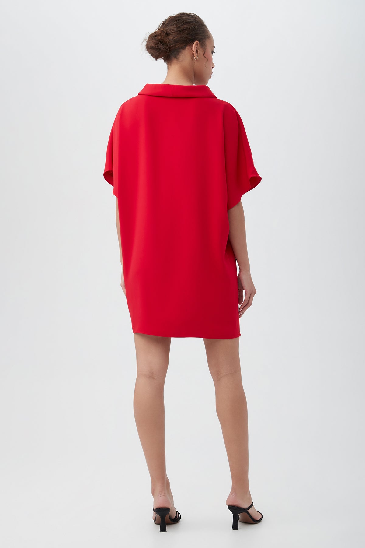 A woman with her hair tied back models the Honeysuckle Short Dolman Sleeve Shift Dressâ€” a loose-fitting, collared red dress with short sleevesâ€”paired with black high-heeled sandals, standing against a plain white background.