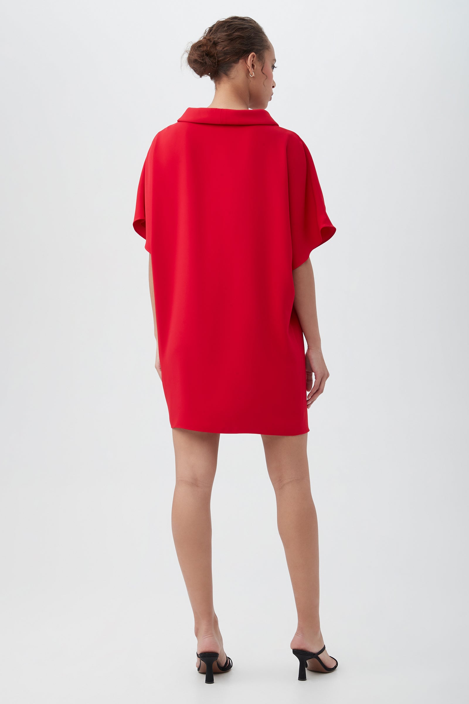 A woman with her hair tied back models the Honeysuckle Short Dolman Sleeve Shift Dress— a loose-fitting, collared red dress with short sleeves—paired with black high-heeled sandals, standing against a plain white background.