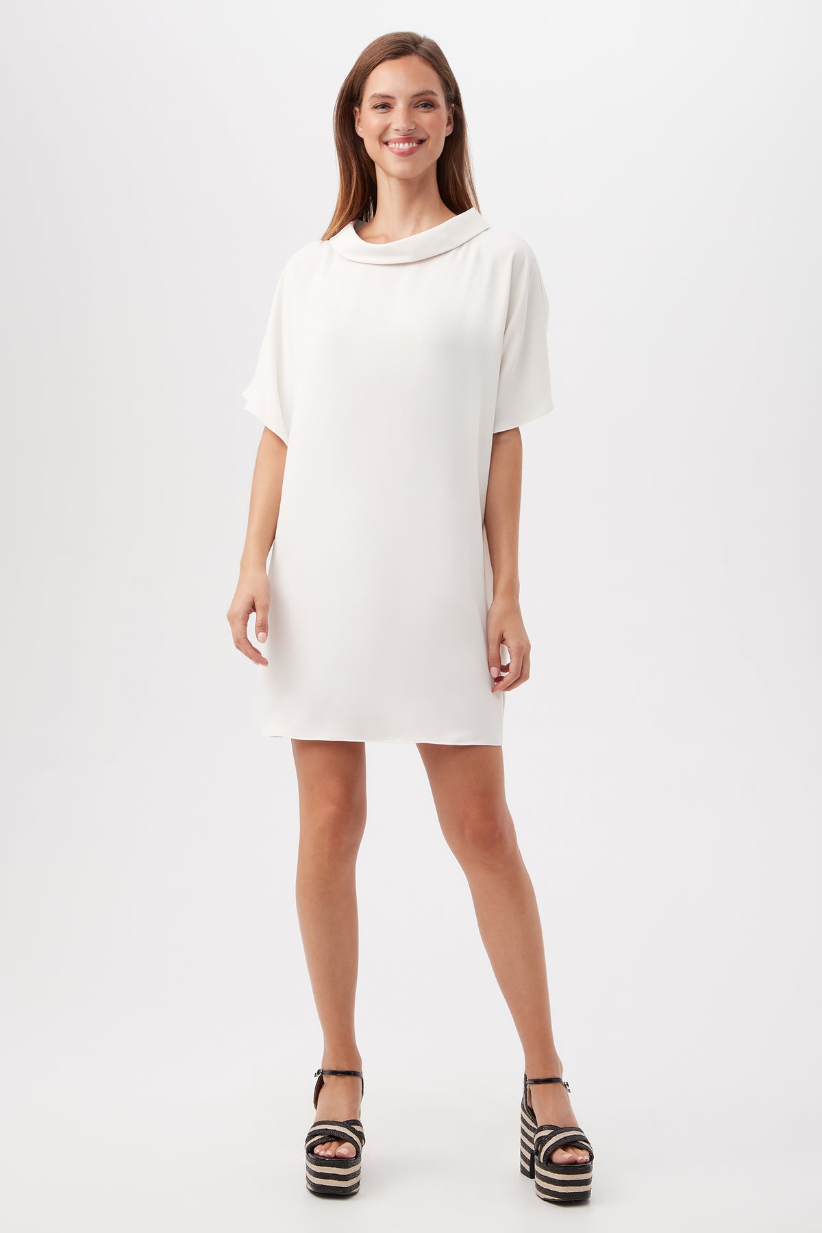 A woman smiles in the Honeysuckle Short Dolman Sleeve Shift Dress in Winter White, paired with black platform sandals, against a plain white background.