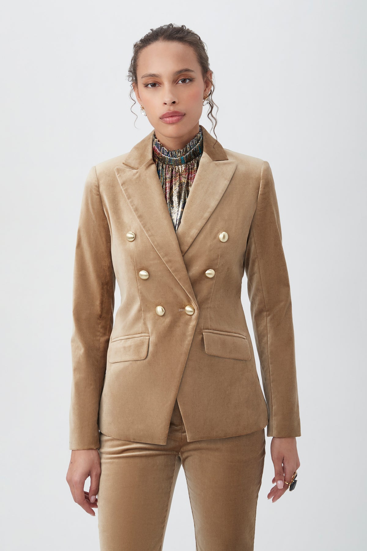A woman stands against a plain background, wearing the Midlands Velveteen Blazer Jacket in gold with matching pants and a patterned high-neck top, looking confidently at the camera.