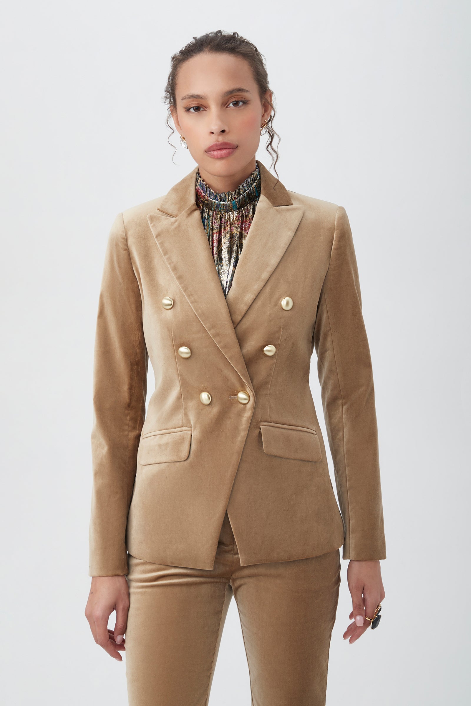 A woman stands against a plain background, wearing the Midlands Velveteen Blazer Jacket in gold with matching pants and a patterned high-neck top, looking confidently at the camera.