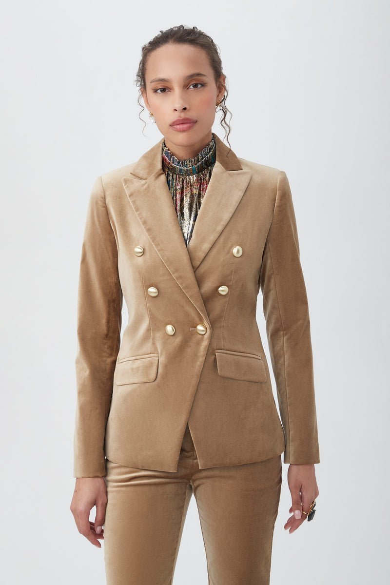 A woman stands against a plain background, wearing the Midlands Velveteen Blazer Jacket in gold with matching pants and a patterned high-neck top, looking confidently at the camera.