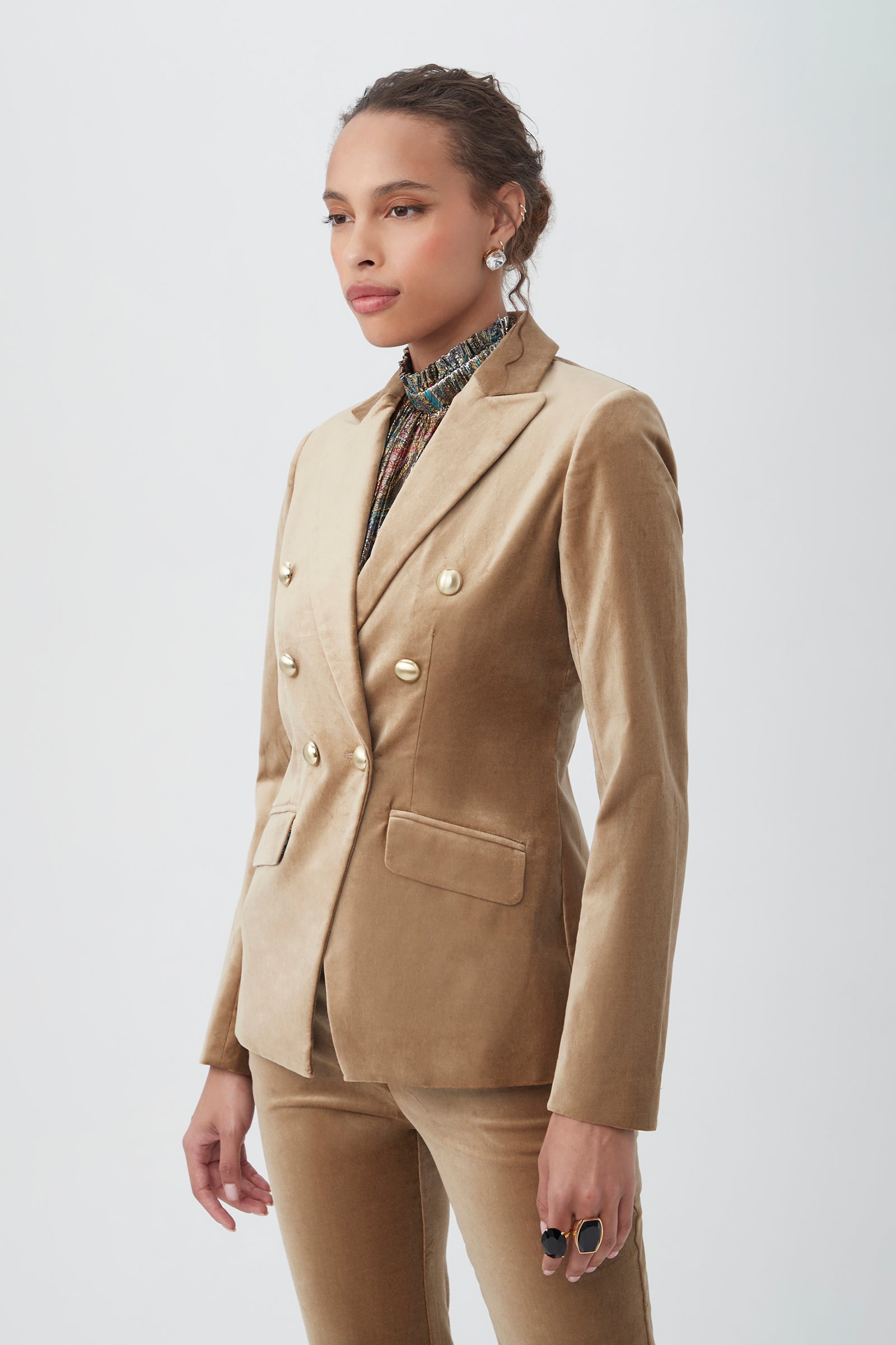A woman stands against a plain background wearing the tan Midlands Velveteen Blazer Jacket and matching pants, styled with statement rings, stud earrings, and a high-neck patterned blouse.