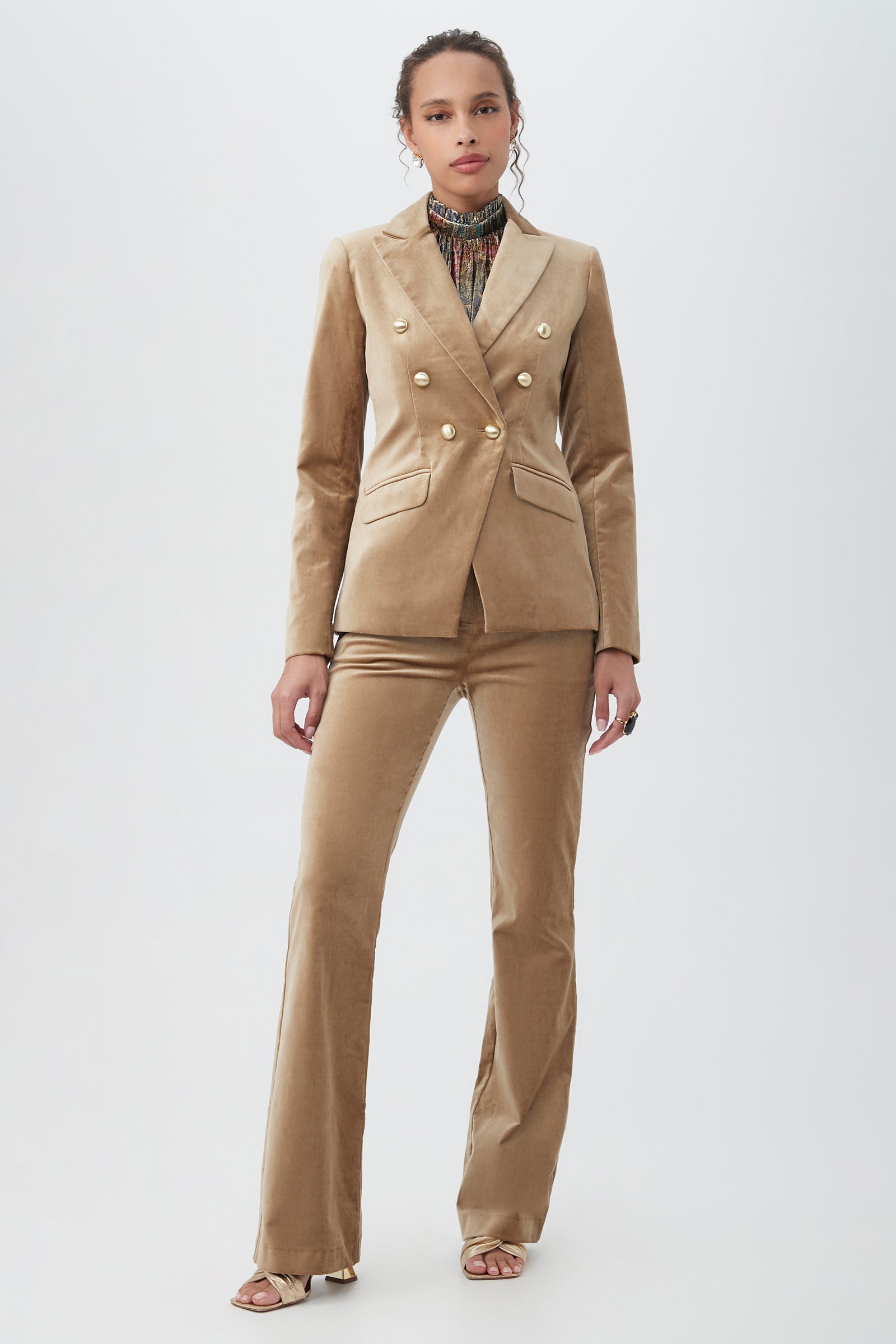 A woman stands against a plain background wearing the Midlands Velveteen Blazer Jacket in tan with gold buttons, paired with matching flared pants, a printed turtleneck, and open-toed heels.