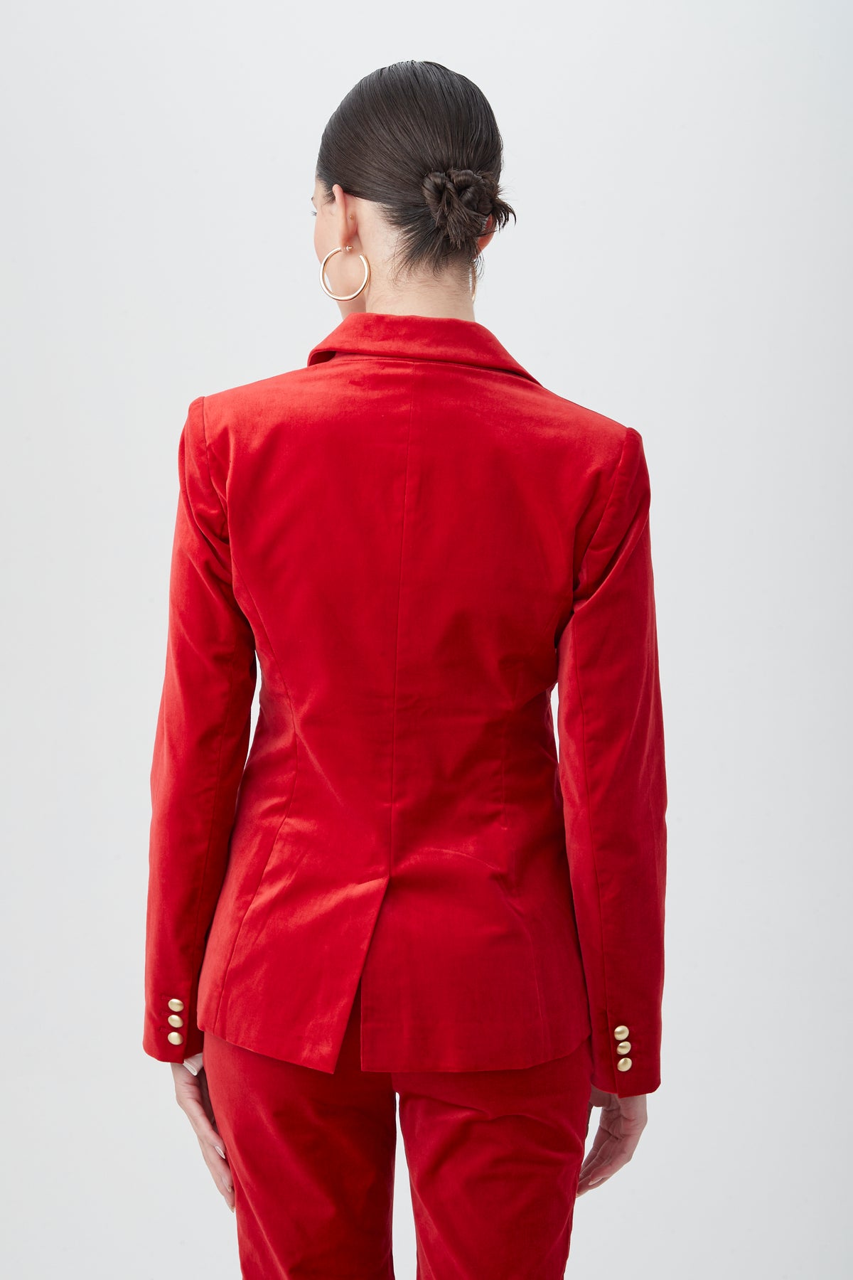 A person with dark hair in a low bun is photographed from behind against a plain light background, wearing the Midlands Velveteen Blazer Jacket and large hoop earrings.