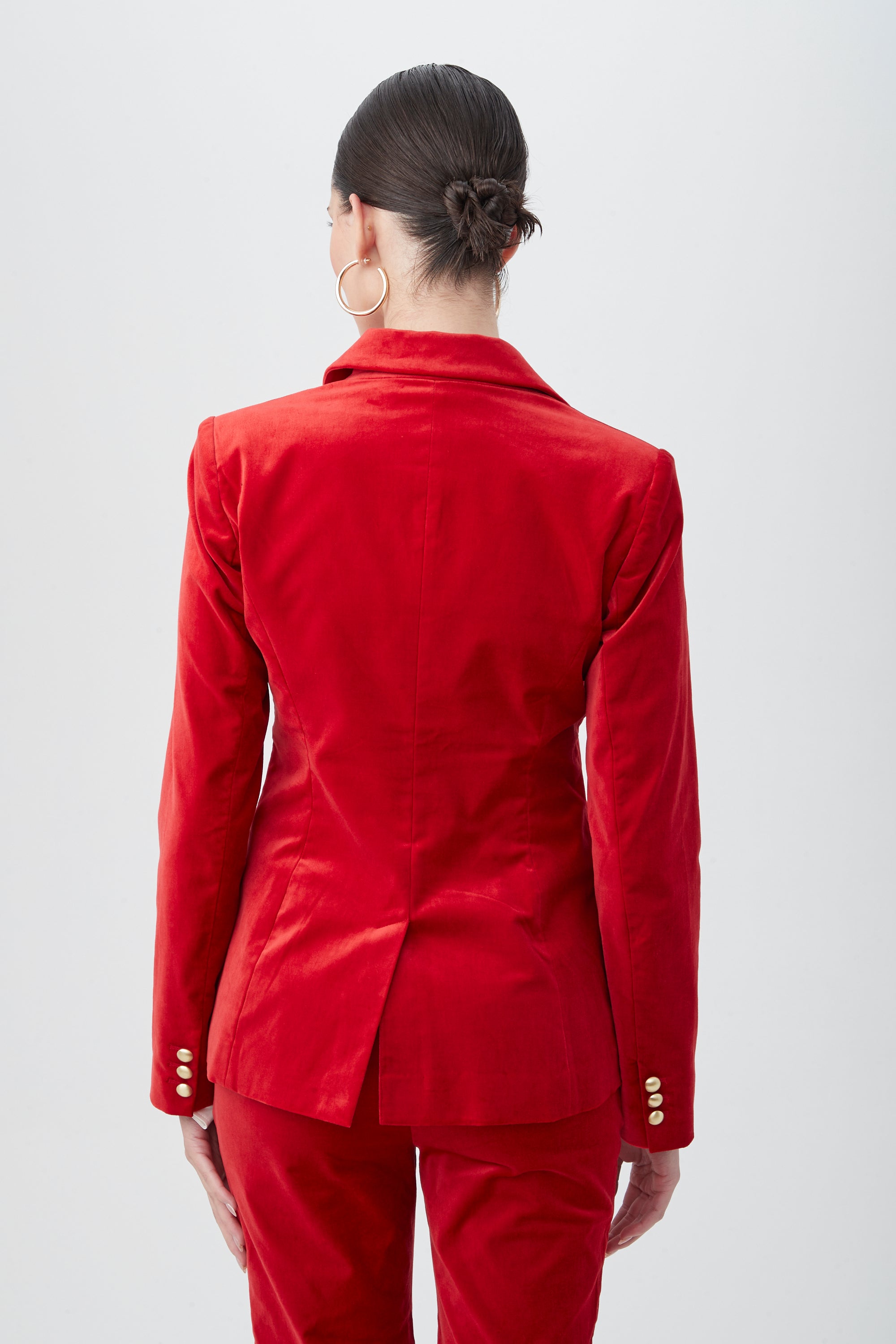 A person with dark hair in a low bun is photographed from behind against a plain light background, wearing the Midlands Velveteen Blazer Jacket and large hoop earrings.