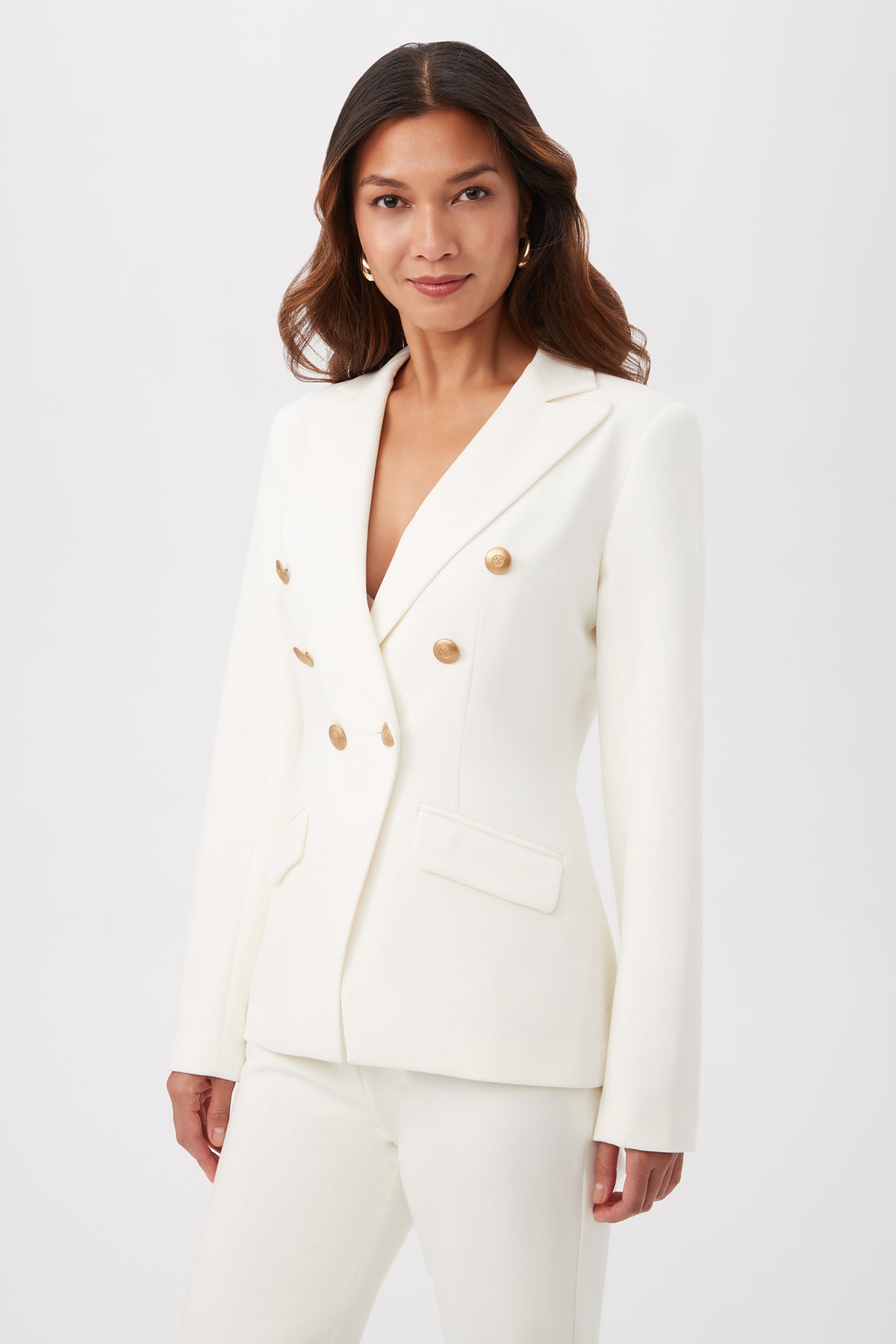 A woman wearing the Midlands Luxe Drape Stretch Blazer Jacket in white with gold buttons and matching pants stands against a plain light background, looking at the camera with a slight smile.