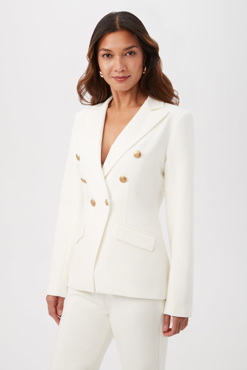A woman wearing the Midlands Luxe Drape Stretch Blazer Jacket in white with gold buttons and matching pants stands against a plain light background, looking at the camera with a slight smile.